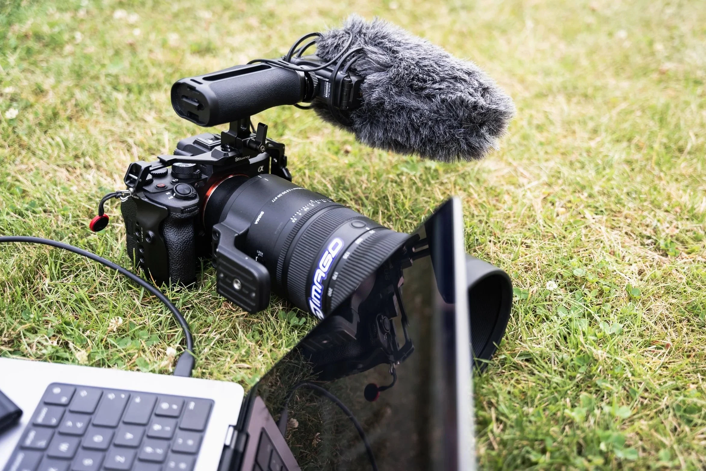 A professional video camera with a large microphone attached, positioned outdoors on grass, with a laptop and other equipment nearby.