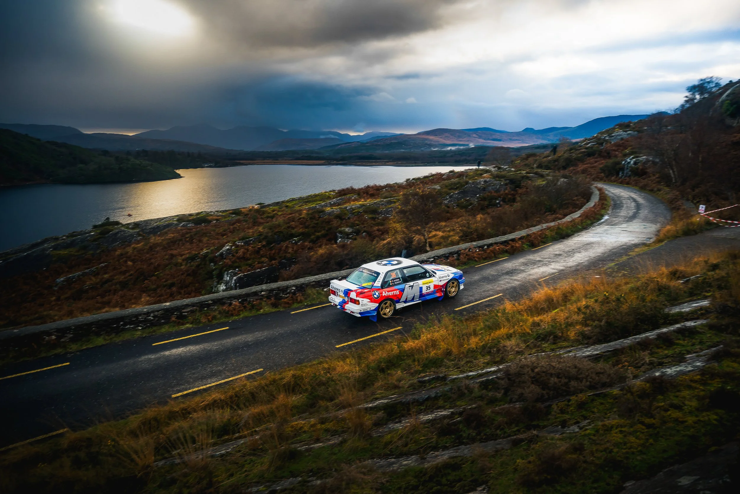 A rally car on a winding mountain road near a lake with dark, cloudy sky in the background.