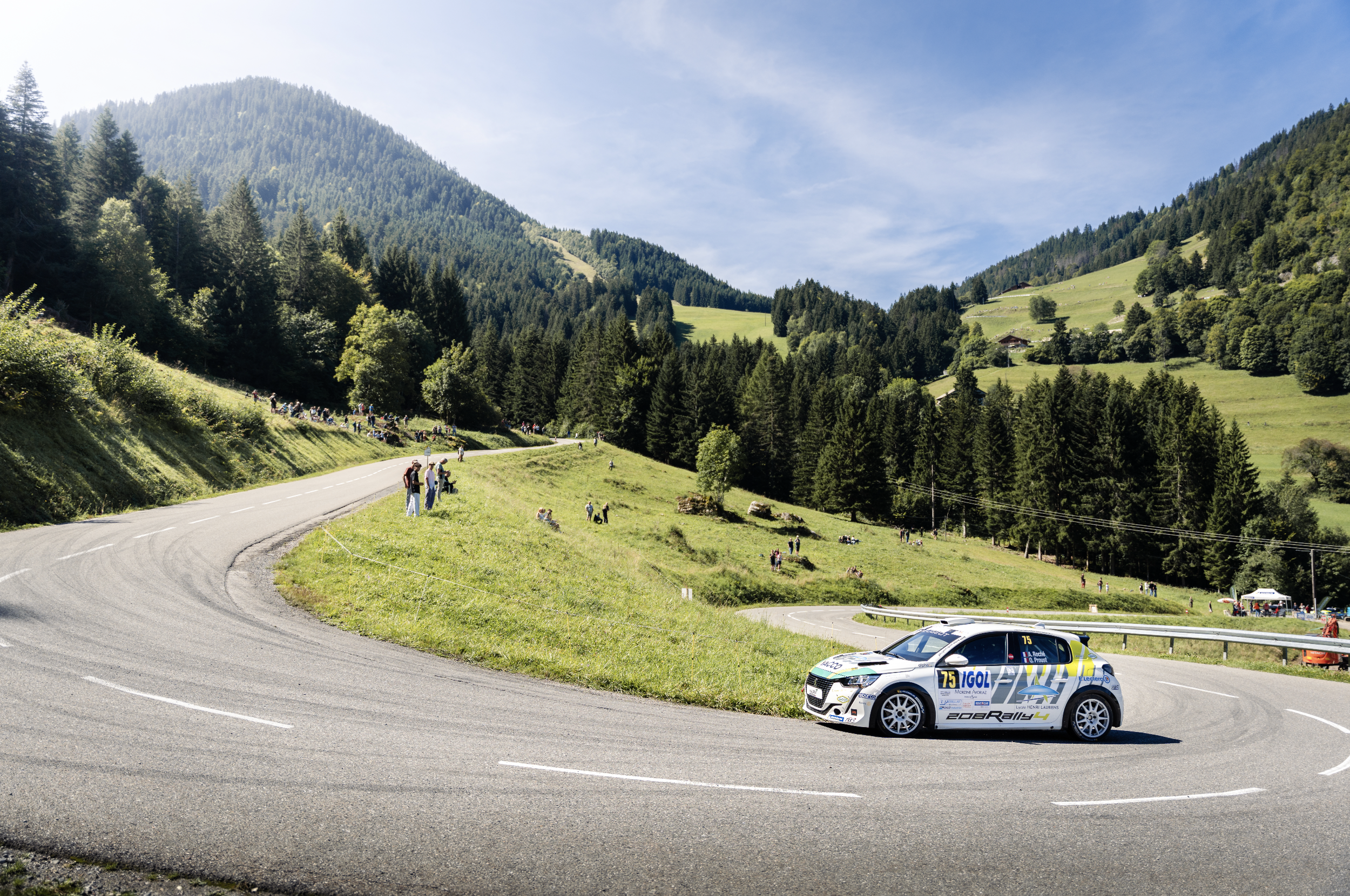 A race car on a winding mountain road with spectators along the side and in the background, lush green hills and forest under a partly cloudy sky.