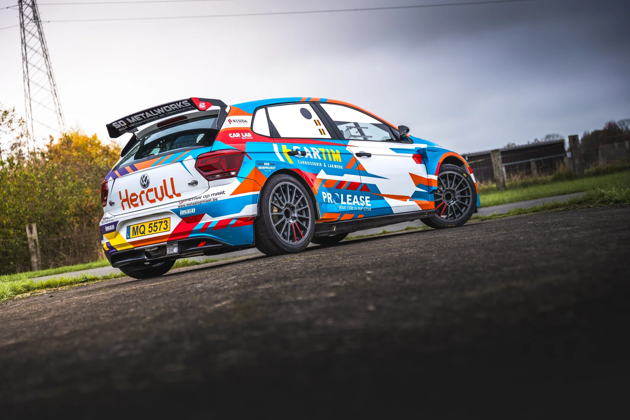 A rally car with colorful sponsor decals parked on a road with a fence and trees in the background under a cloudy sky.