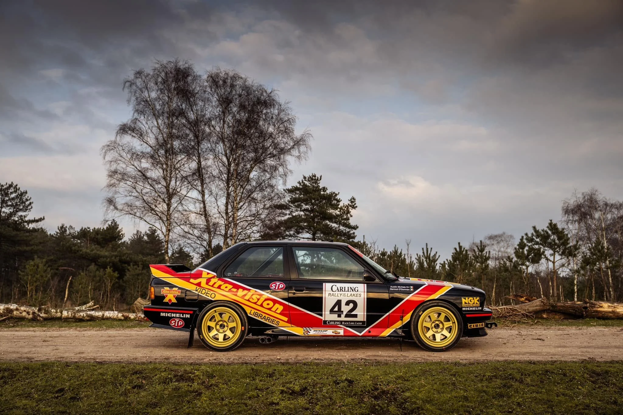 A black rally car with yellow wheels, decorated with racing decals and sponsor logos, parked on a dirt road with trees and a cloudy sky in the background.