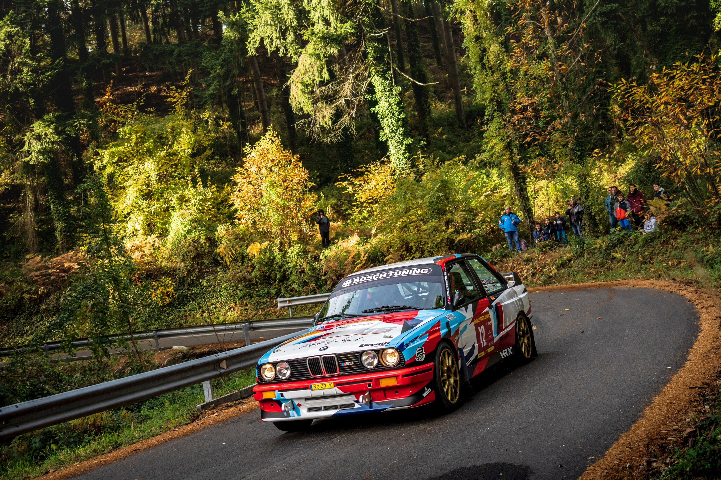 A race car navigating a sharp turn on a forested mountain road during a rally event, with spectators watching from the side.