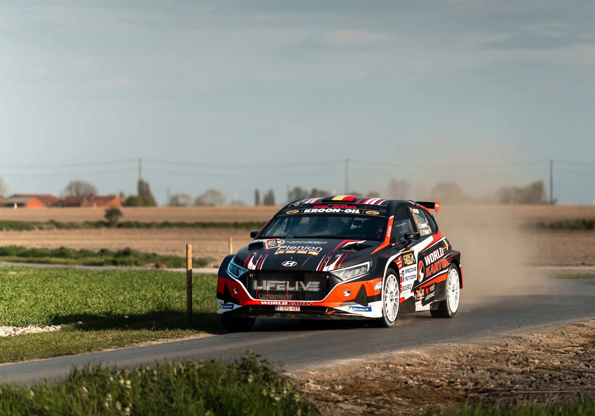 Race car on a dirt track with grass in the foreground and a rural landscape in the background.