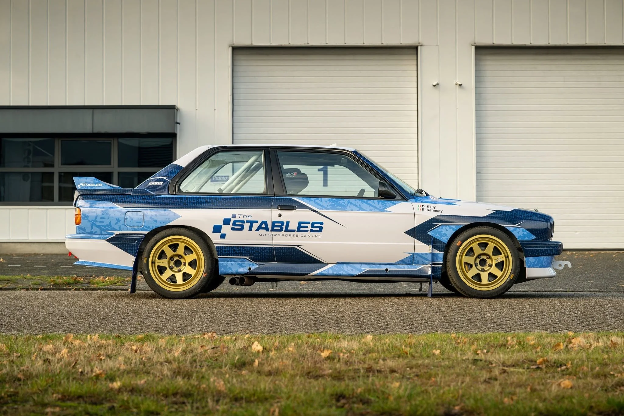 A blue and white race car with yellow wheels parked outside a large industrial building with closed garage doors.