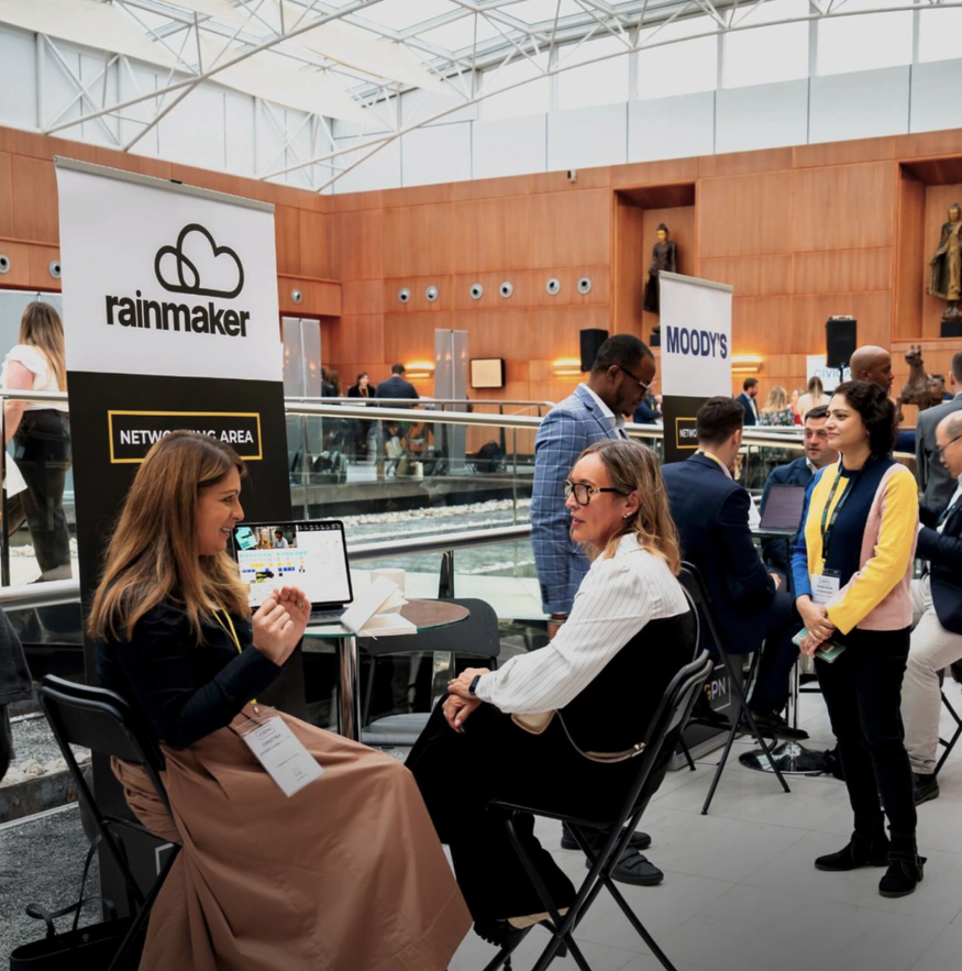 Business conference with people engaging at booths labeled 'rainmaker' and 'MOODY'S' inside a large, modern building.