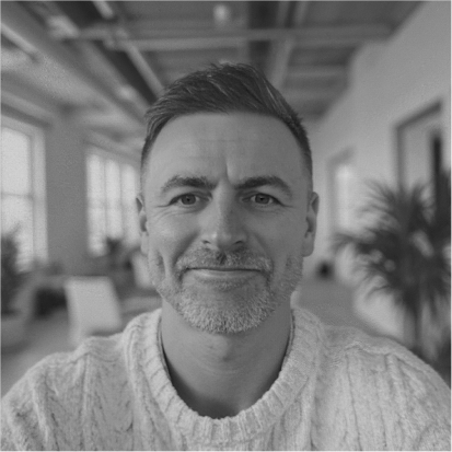 Black-and-white photo of a man with short hair and a beard, smiling, indoors with windows and plants in the background.