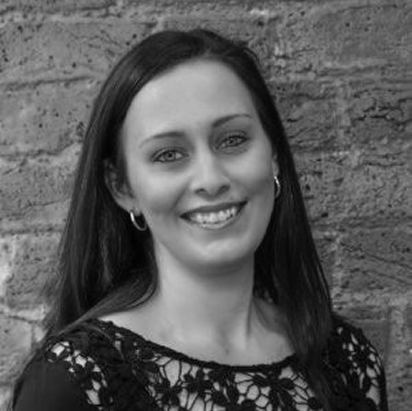 A woman with dark hair smiling, standing in front of a brick wall, wearing a black lace top and hoop earrings.