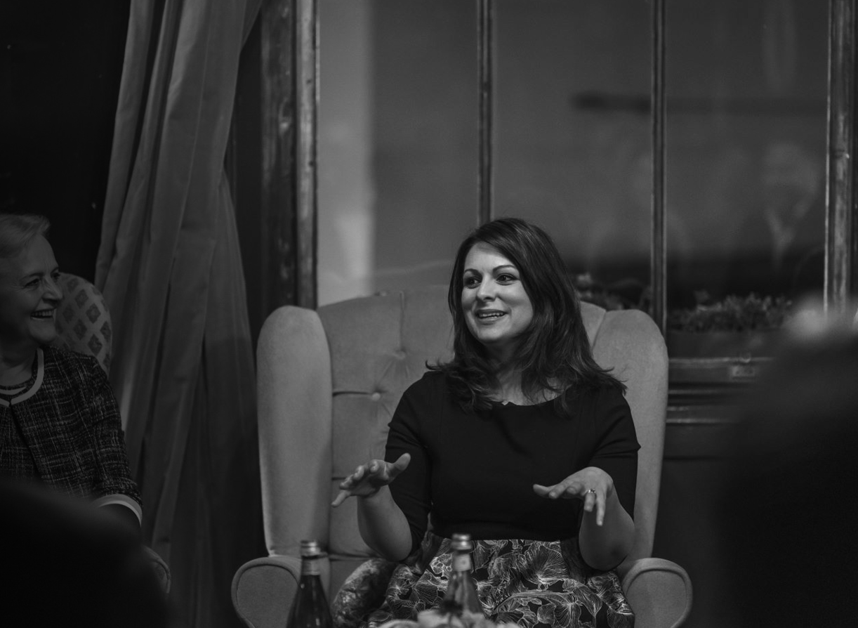 A woman with shoulder-length dark hair, wearing a black top and patterned skirt, sitting on an armchair, gesturing with her hands, engaged in conversation with a woman on her left, in a room with a wooden wall and a window in the background.