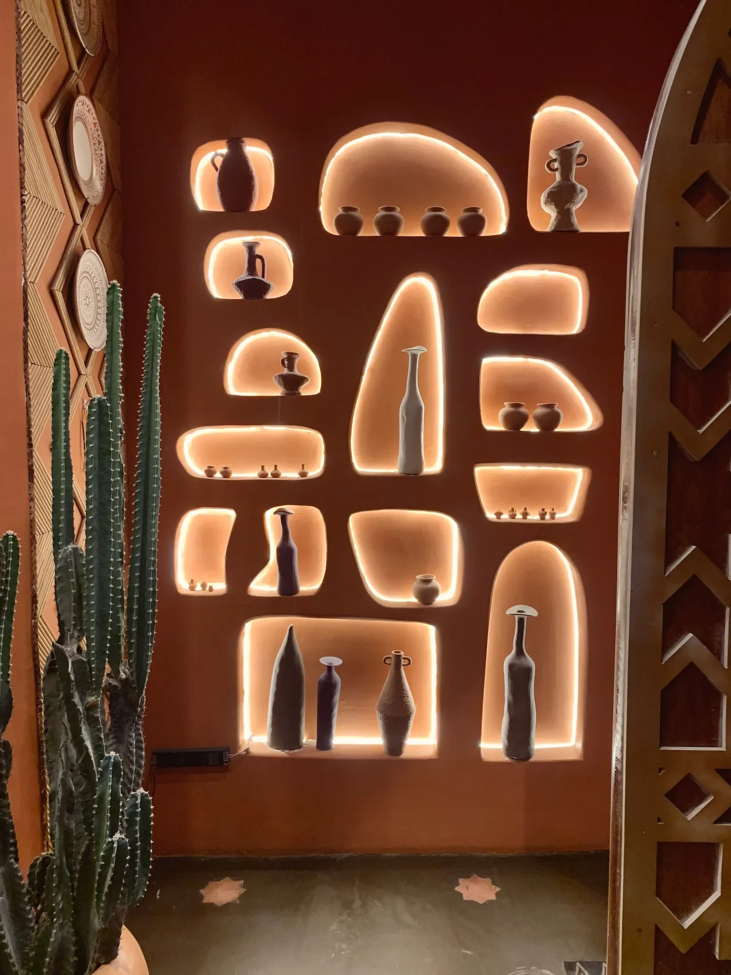 Objets en céramique sur des niches éclairées à l'arrière, décor minimaliste avec cactus à gauche et sol en bois.