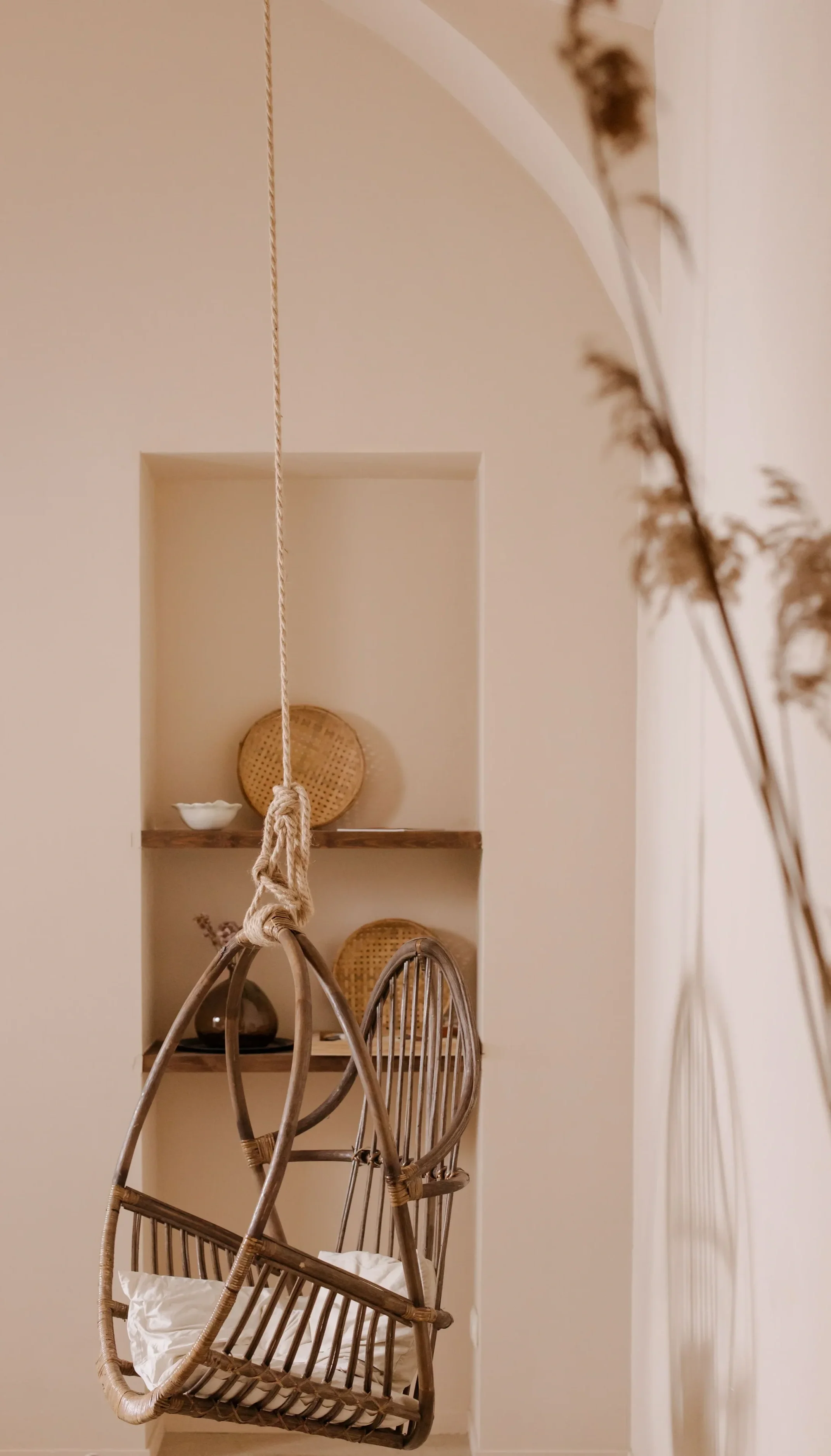 Une balançoire en rotin suspendue par une corde, dans une pièce aux murs beige clair. En arrière-plan, une étagère encastrée avec des éléments décoratifs en rotin, et une partie d'une plante sèche au premier plan droit.