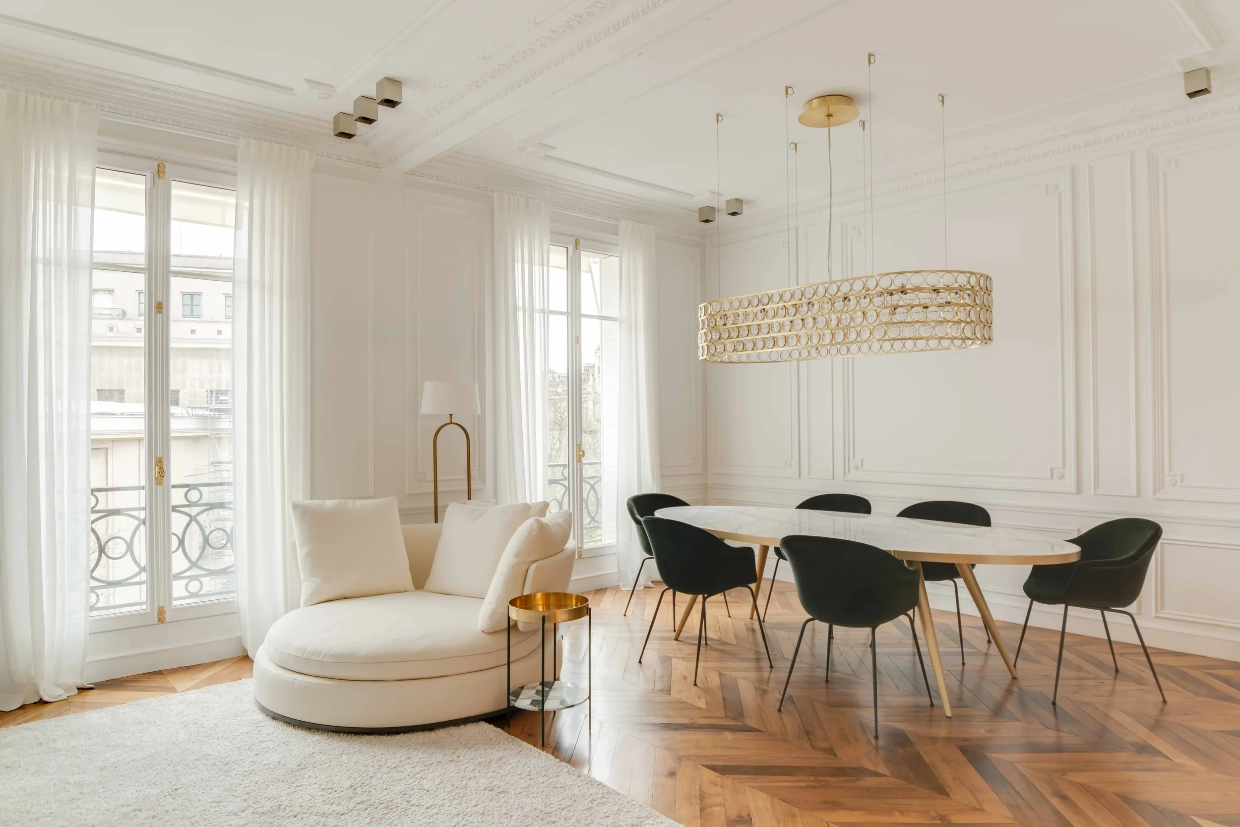 Salon lumineux avec grandes fenêtres, canapé blanc, lampe dorée, table ronde, chaises noires, lustre moderne et murs avec moulures en bois.