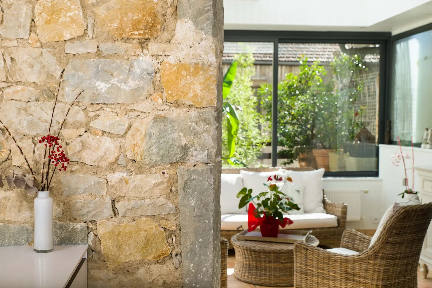Salon type véranda avec canapé blanc et osier, fauteuils en rotin, table basse, grande verrière avec vue sur une terrasse végétalisée extérieure.