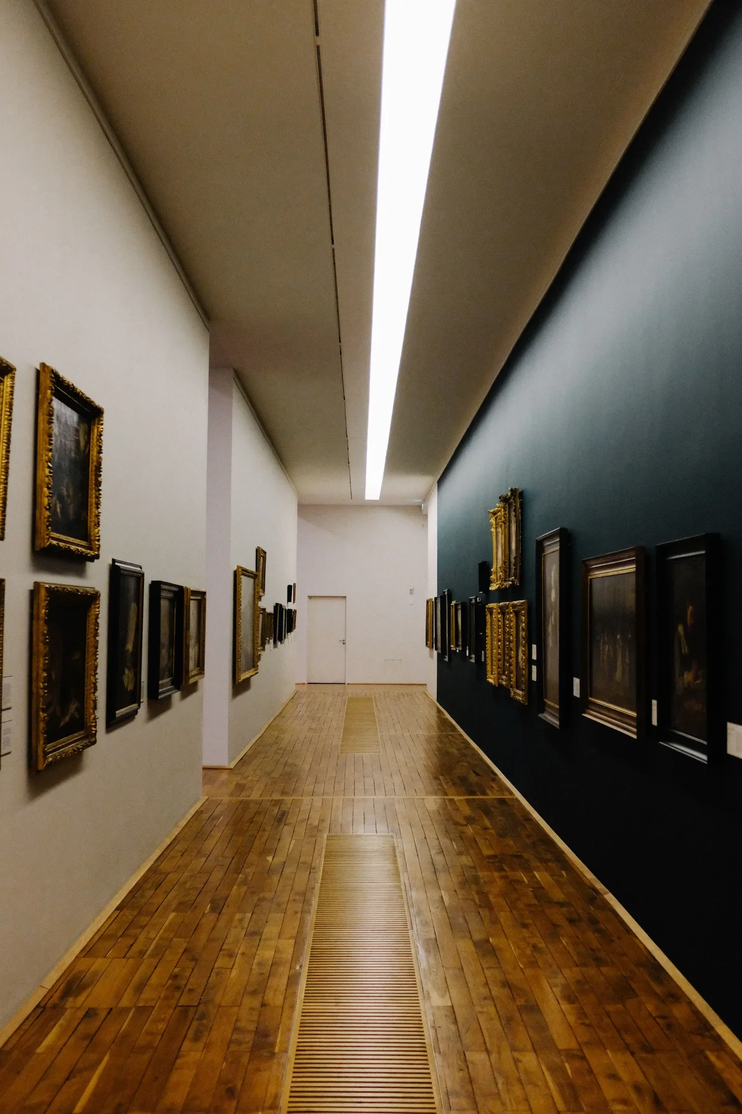 Un couloir de musée avec un jeu de contraste un mur blanc et un mur bleu pétrole ornés de tableaux encadrés dorés. Le sol est en parquet de bois clair et au centre. La lumière provient d’un plafonnier long, suspendu au plafond.