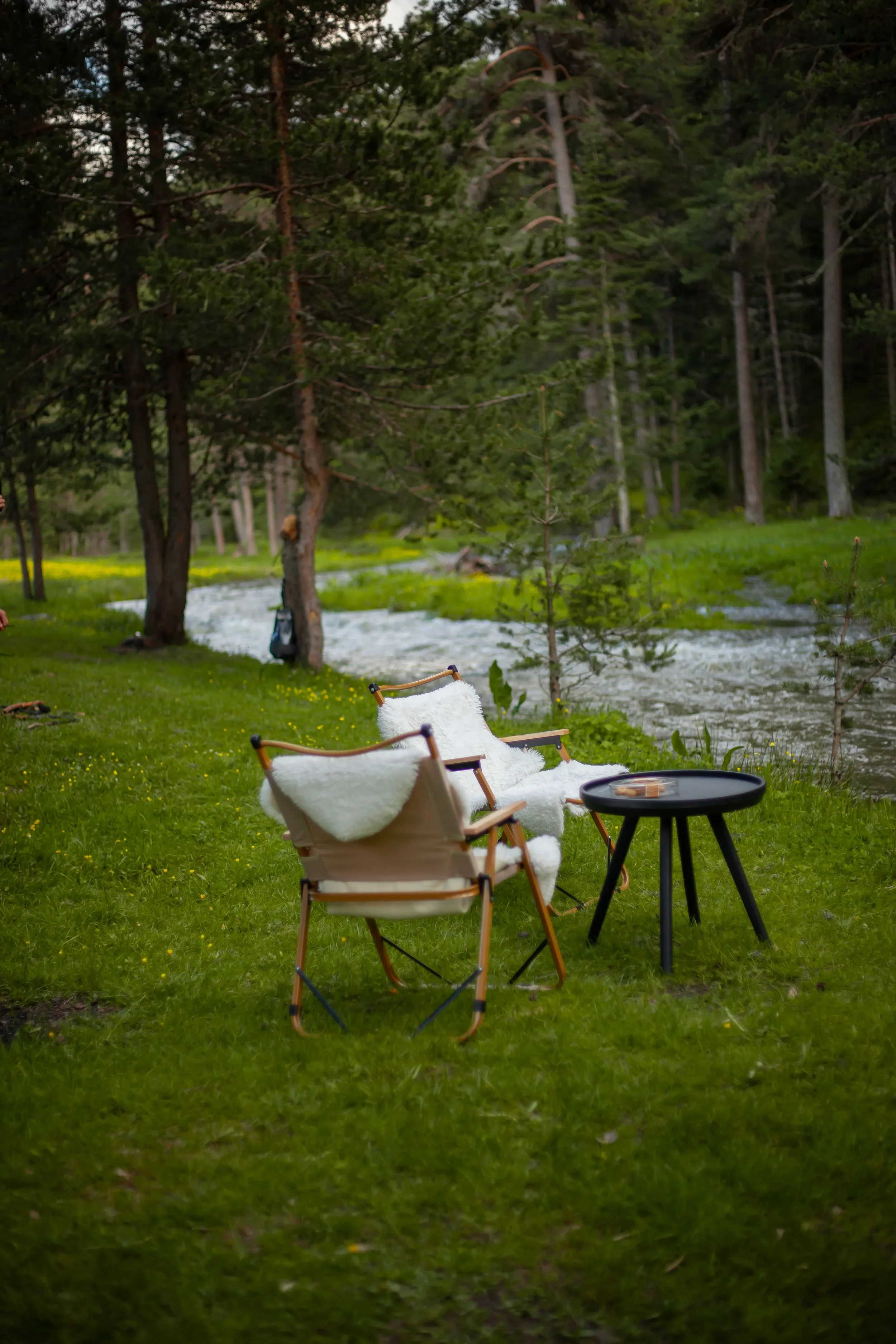 Deux fauteuils en bois avec des coussins blancs en peluche sont placés sur une pelouse verte à côté d'une petite table noire ronde, dans un environnement boisé près d'une rivière.