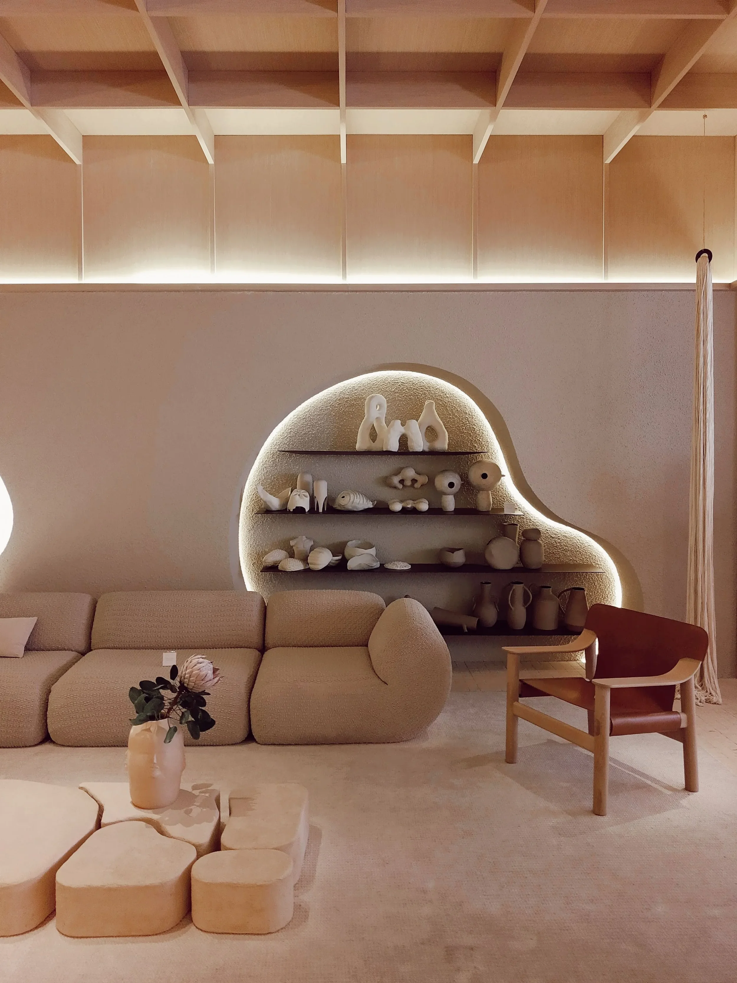 Un salon avec un canapé beige, une table basse en forme de branche avec un vase contenant des fleurs, une étagère en forme d'arc avec des objets en céramique blanche, et un fauteuil en bois avec un coussin beige. La pièce est décorée dans un style minimaliste avec une ambiance chaleureuse.
