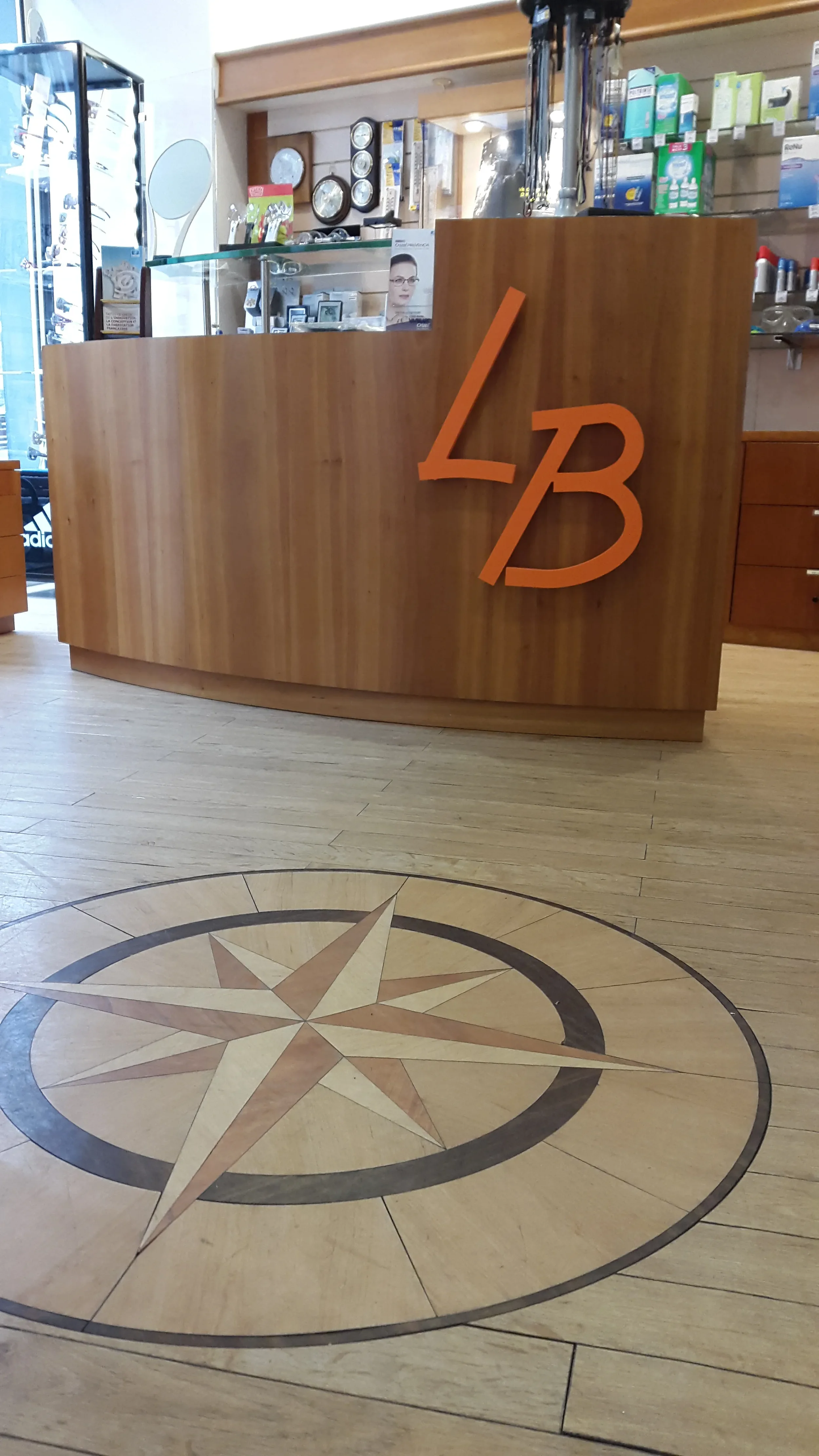 Un comptoir d’accueil sur mesure réalisé en bois avec les initiales "LB" fixées en applique, pour un commerce d'optique en centre ville. Élégant sol en parquet avec un motif de rose des vents incrusté.
