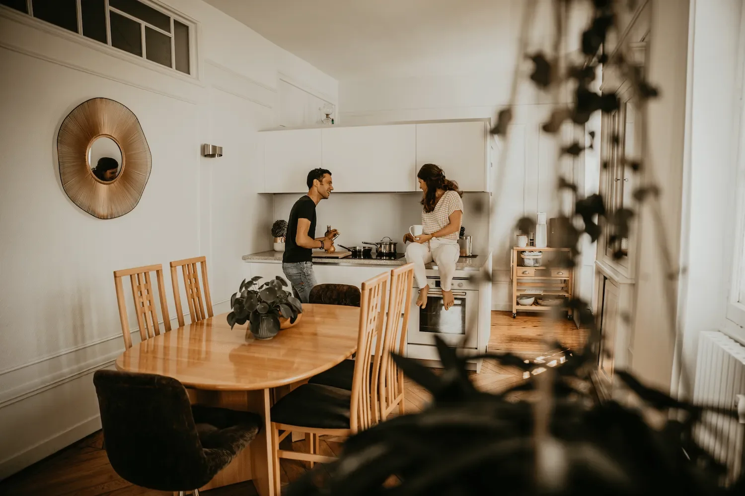 Deux personnes dans la cuisine, un homme et une femme, discutent. La femme est assise sur le comptoir et l'homme se tient à côté. La cuisine est moderne, avec une table en bois et des chaises, et une décoration intérieure chaleureuse.