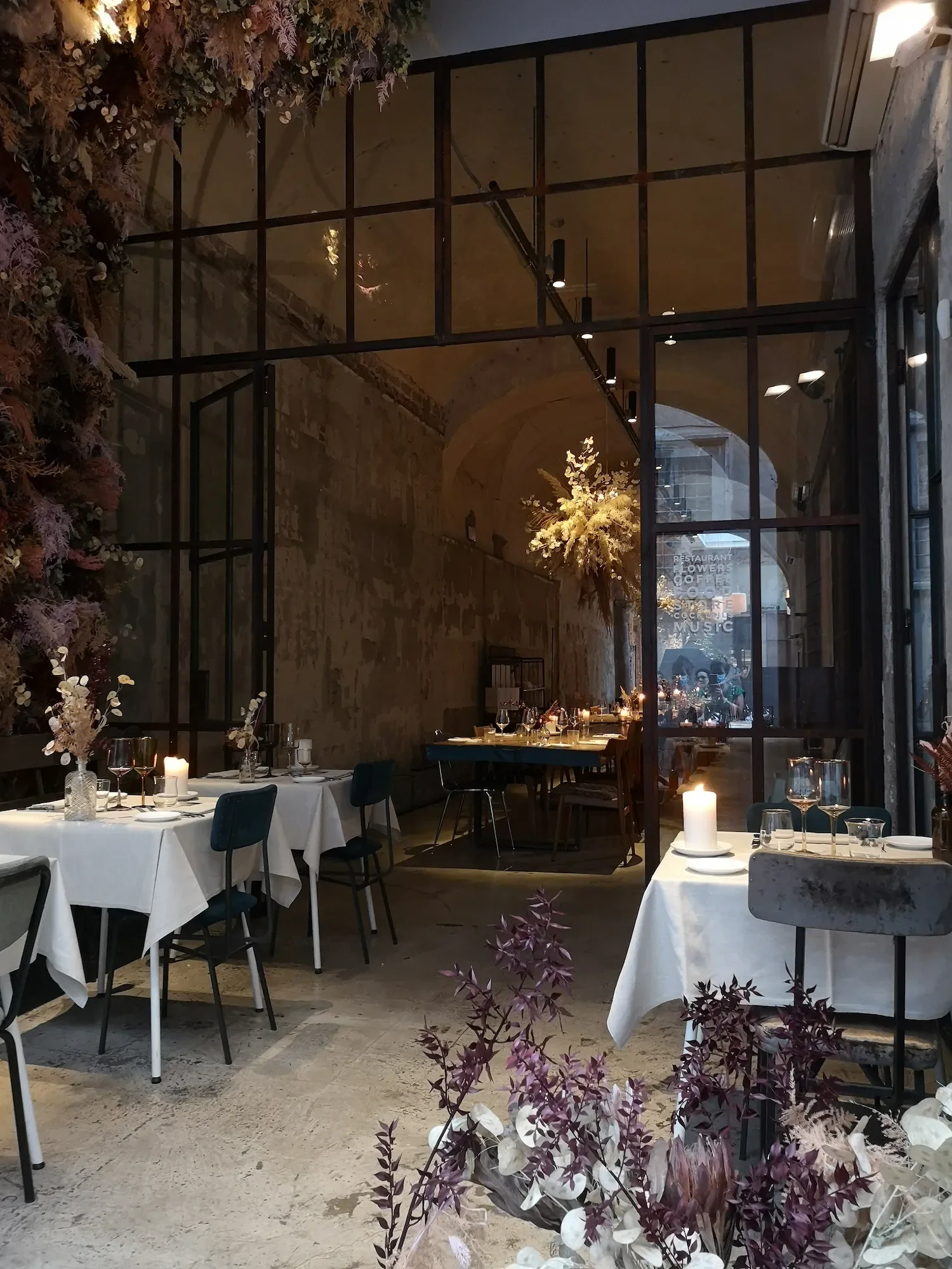 Intérieur d'un restaurant florentin élégant avec des tables dressées, des bougies et des fleurs séchées, un sol en béton brut, des murs en pierre, vue à travers une magnifique verrière.