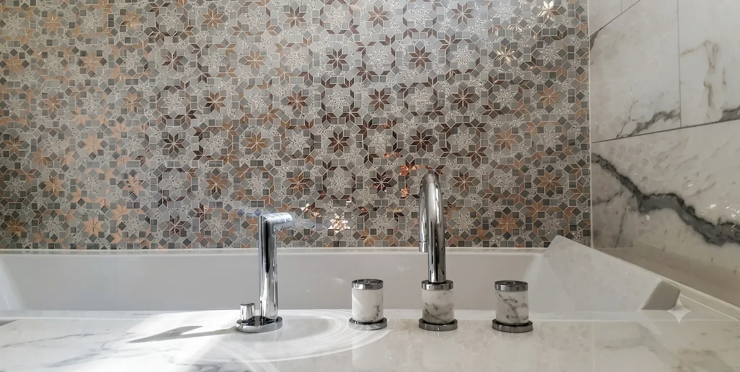 Mosaïque sur mur de salle de bain avec un décor de carreaux type zellige. Robinetterie en marbre blanc de carrare encastrée sur la baignoire. Création sur mesure.