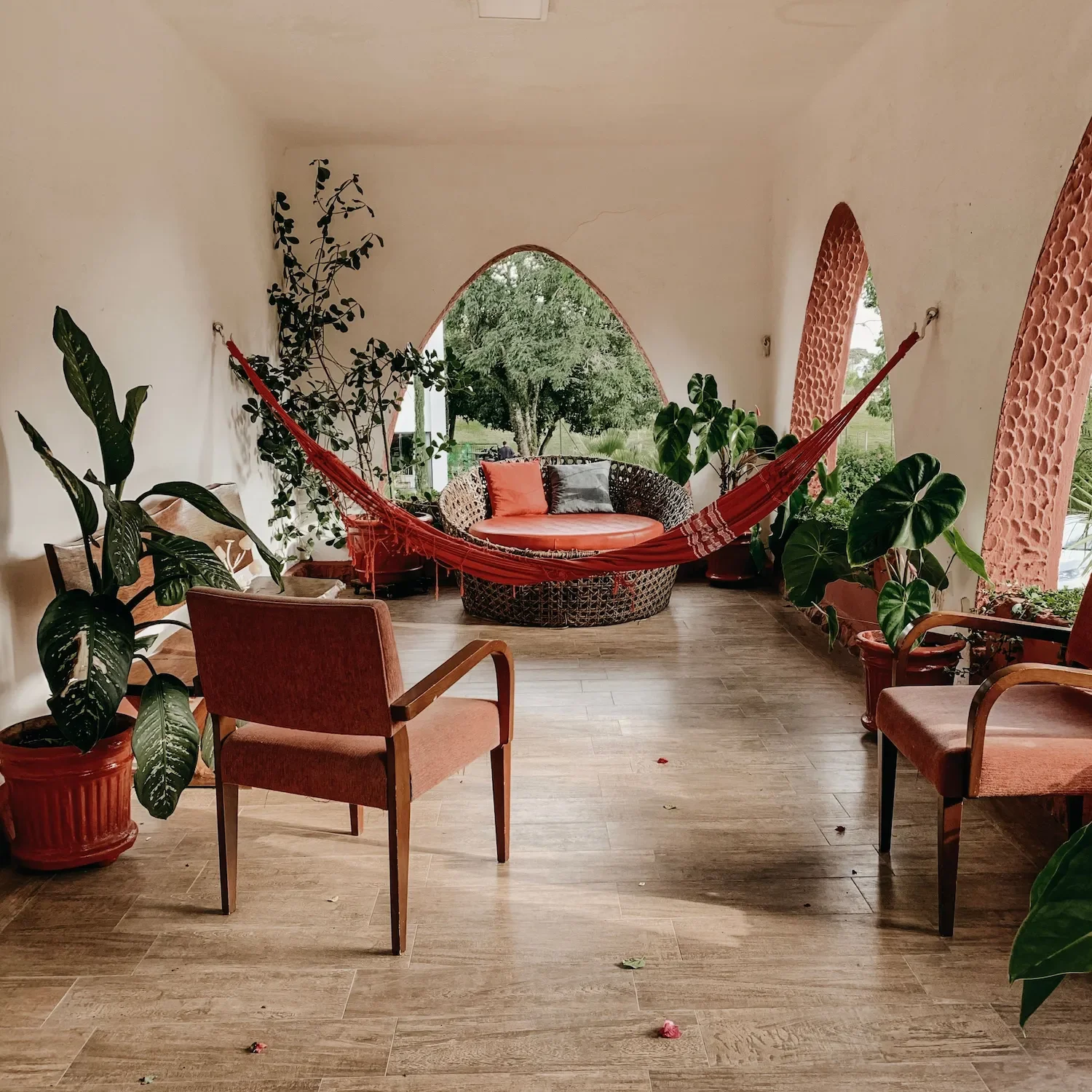 Une véranda avec plusieurs chaises et canapés en bois et tissu, décorée de nombreuses plantes vertes en pot, un hamac rouge suspendu au plafond, avec un mur blanc à l'arrière et une vue de la nature par les arches en arrière-plan.