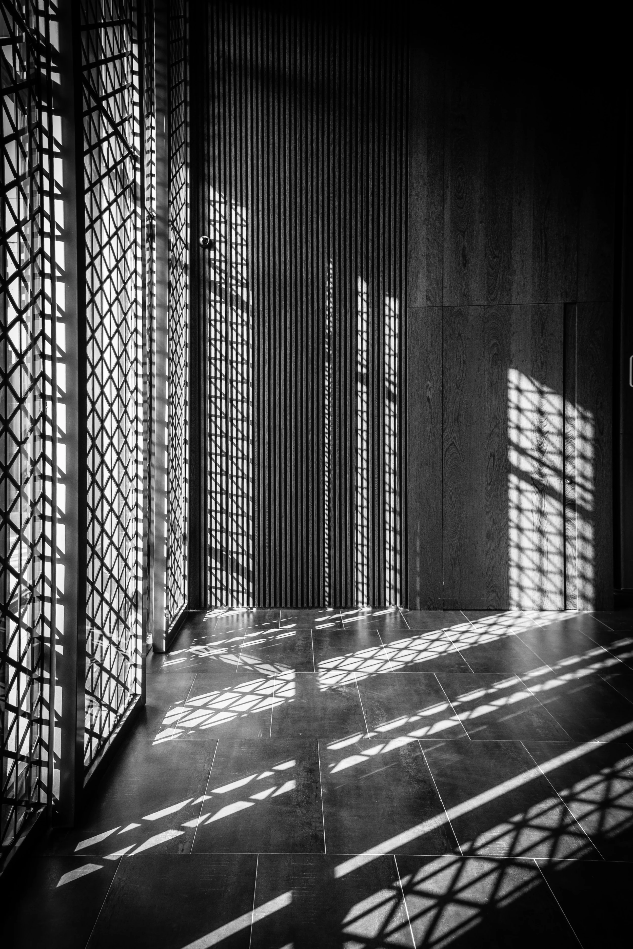 Une pièce sombre avec des murs en bois et des fenêtres en treillis avec des ombres de lumière qui créent des motifs au sol.
