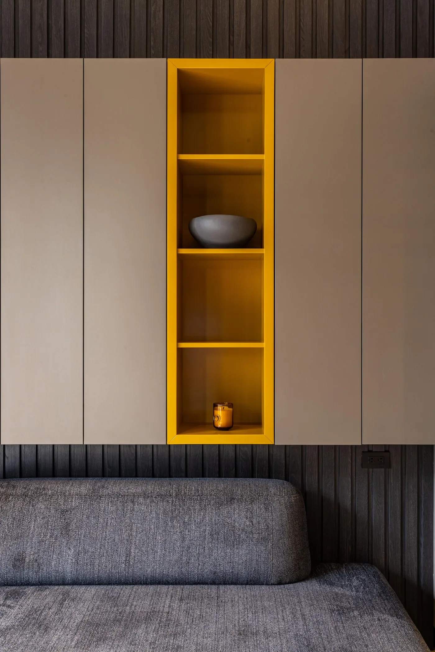 Agencement sur mesure. Une niche jaune centrale avec des rangements pleins en panneaux beige, au-dessus d'un canapé gris.