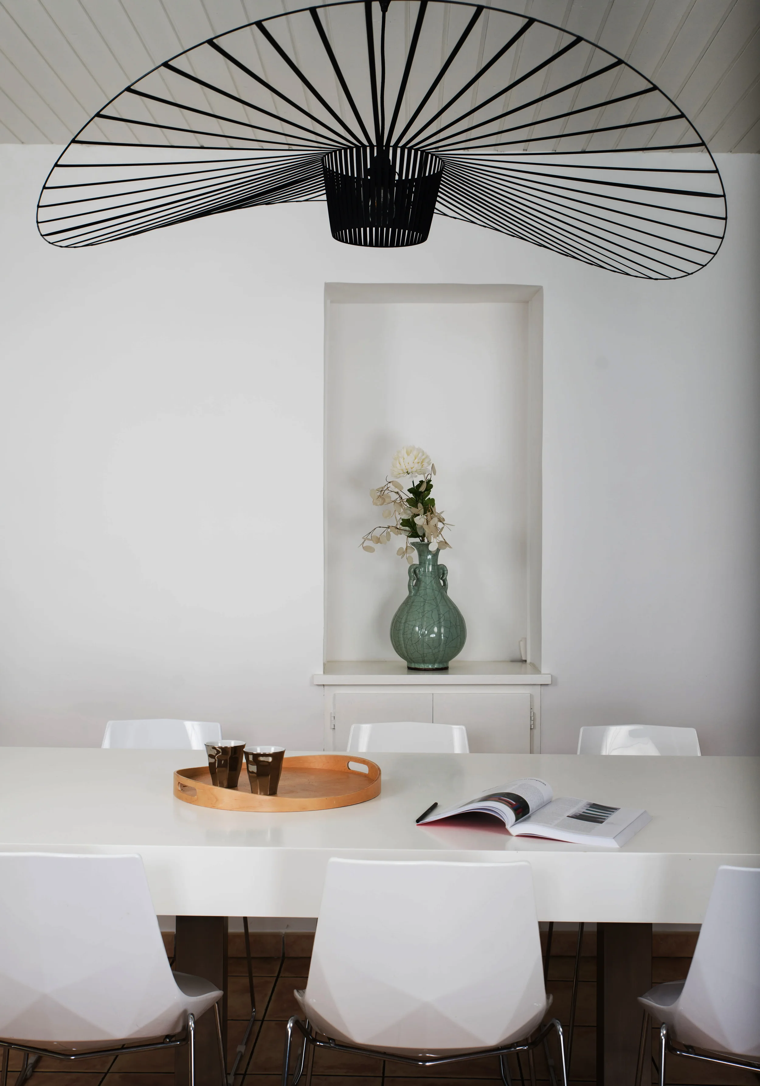 Une salle à manger moderne, épurée et lumineuse. Un mur blanc en arrière plan avec une niche intégrée contenant un vase en céramique émaillé. Le mythique Vertigo (luminaire suspendu) pour éclairer la table avec ses jeux d'ombres et de lumière.