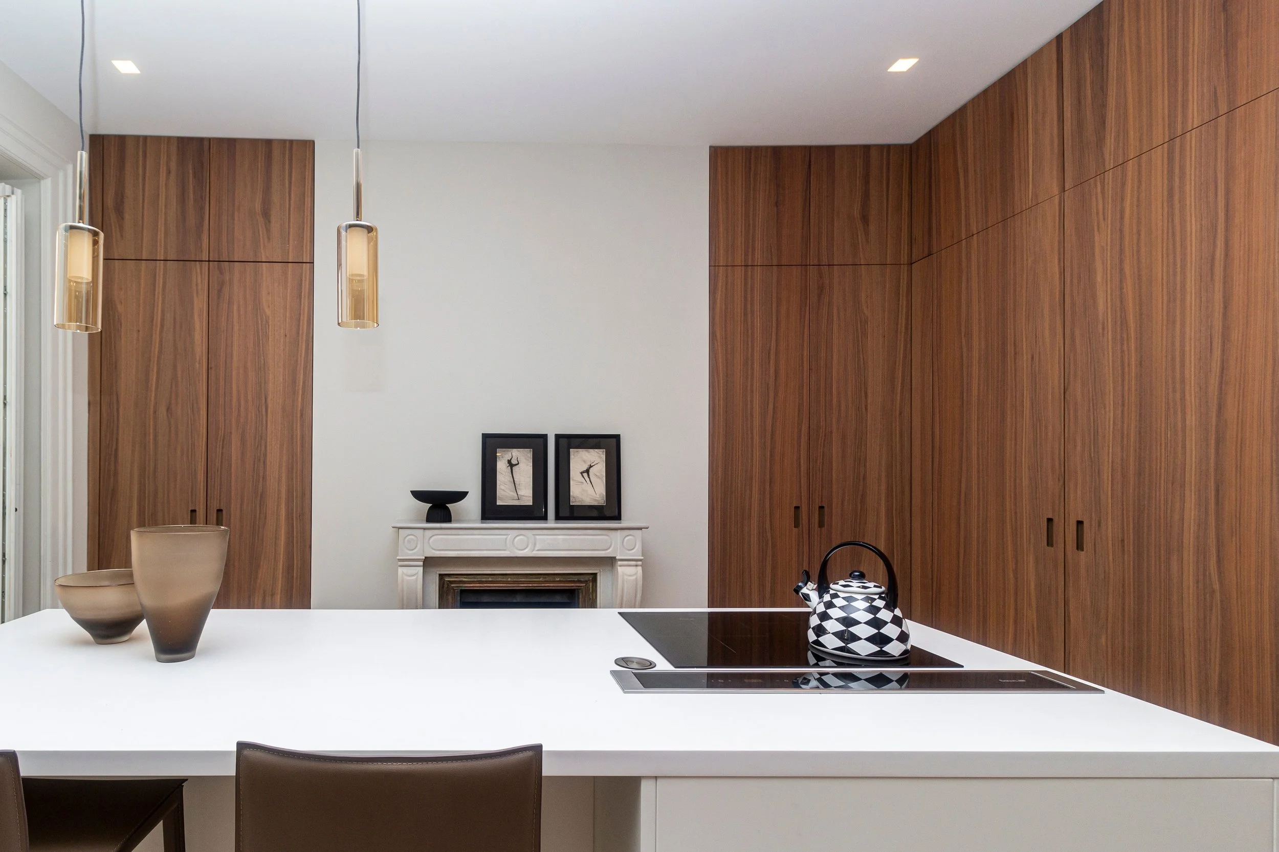 Une cuisine lumineuse et moderne avec un plan de travail en marbre blanc, deux vases en poterie, une plaque de cuisson avec hotte intégrée, un agencement en placage bois pour les colonnes de rangements valorisant une cheminée d'époque au centre.