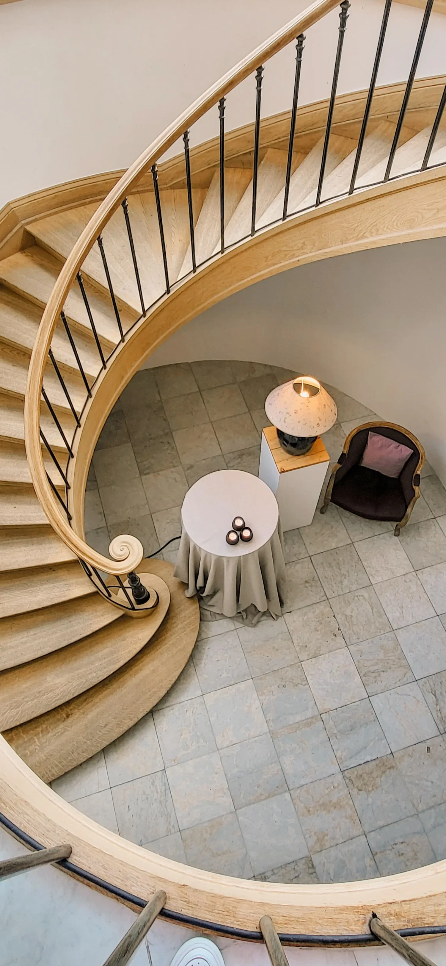 Vue depuis le haut d'une cage d'escalier en forme de spirale, avec un sol en carreaux, un fauteuil, une table avec une nappe, une lampe sur un socle et trois bougies