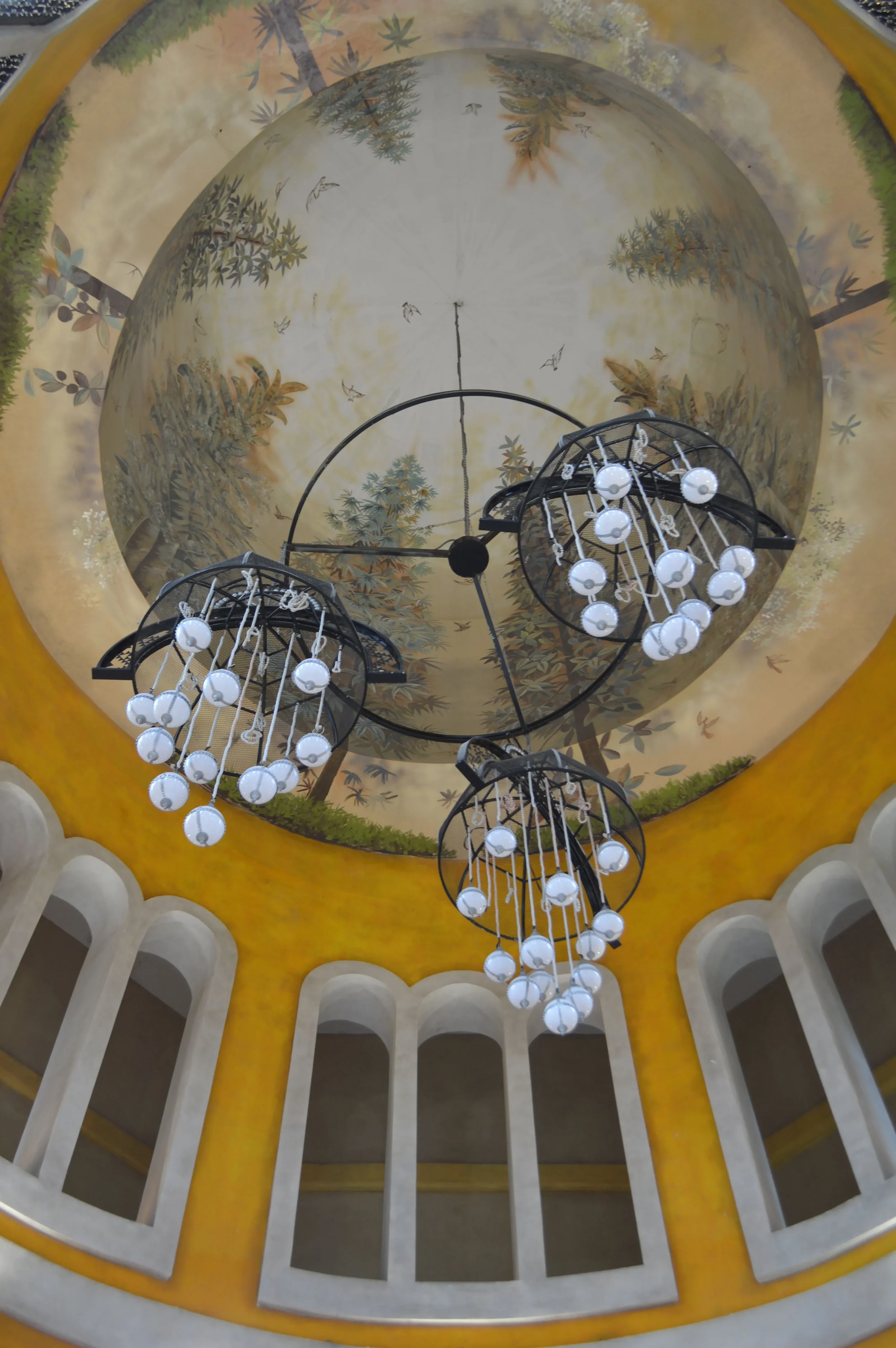 Vue du plafond intérieur avec fresque murale représentant une scène de nature avec des arbres et des oiseaux, ainsi que des lustres suspendus avec des globes lumineux.