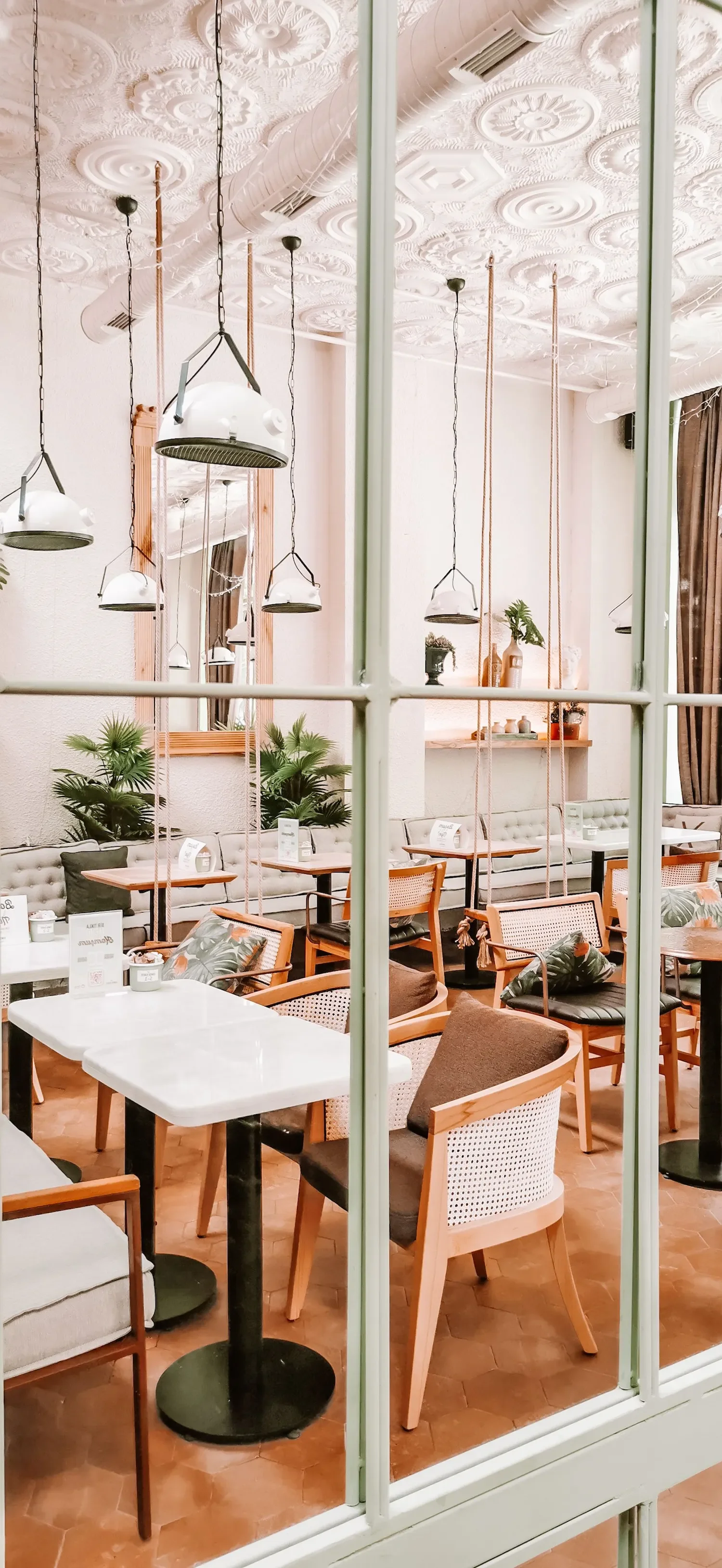Intérieur d'un café ou restaurant avec des tables, chaises en bois, plantes vertes, miroir, suspensions modernes, et décor élégant, vu à travers une fenêtre