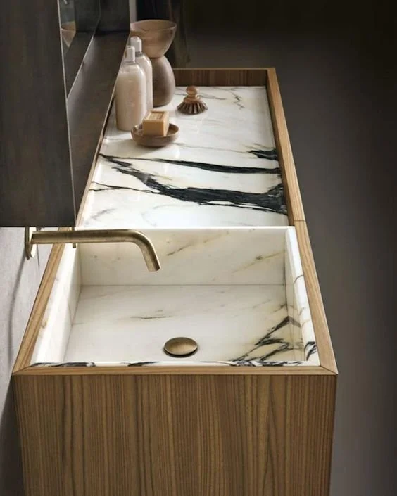 Un lavabo en marbre blanc avec des veines noires, encastré dans une structure en bois naturel, avec un robinet en laiton à gauche. Sur la surface à l'arrière, il y a des jars en céramique, une brosse de nettoyage, et un bol en bois.