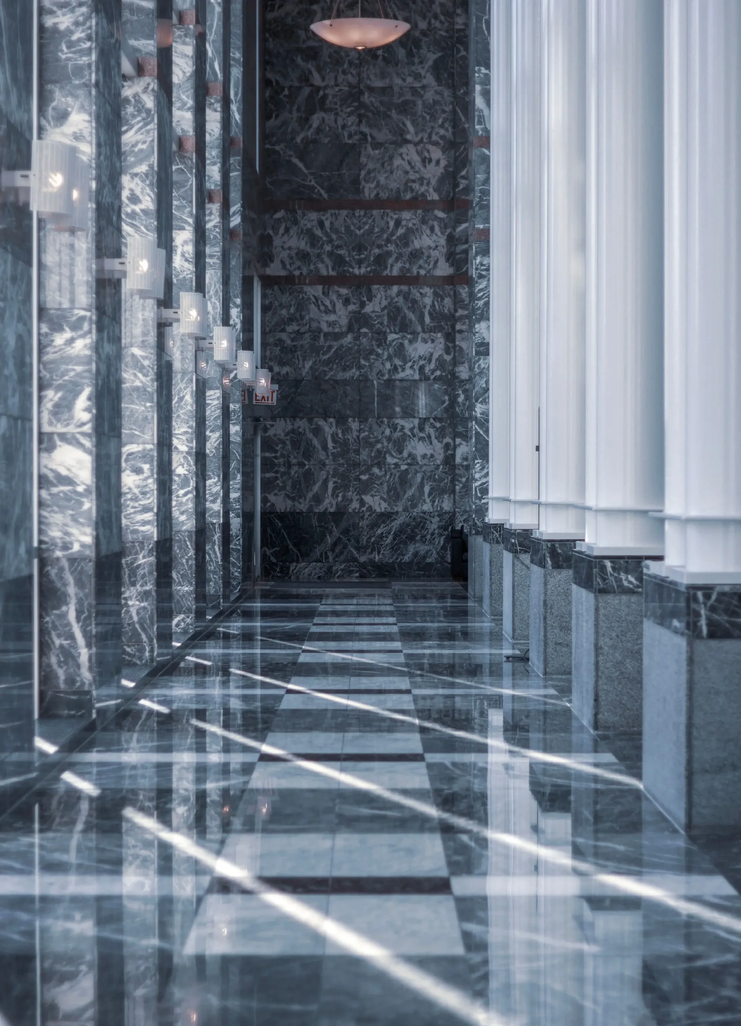 Intérieur minéral d'un bâtiment avec des murs et un sol en marbre sombre veiné, éclairé par des suspensions. Jeux de rythme avec les colonnes, jeux de plein et vide avec le choix du marbre blanc ou gris veiné.