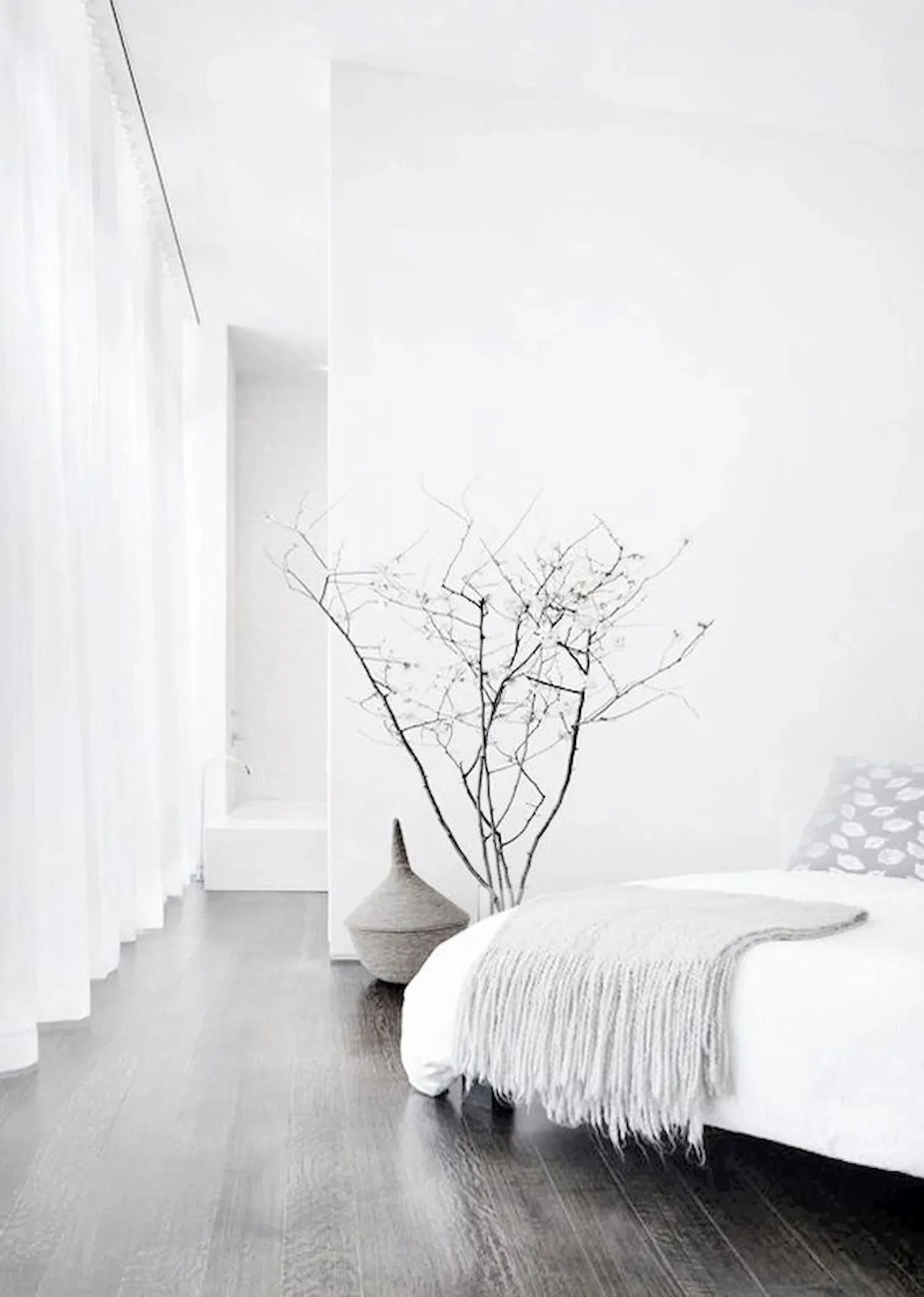 Une chambre minimaliste en noir et blanc avec un lit recouvert d'une couverture texturée, une longue vase en osier avec une branche nue, et un mur blanc avec une arche. La pièce a un sol en bois clair.