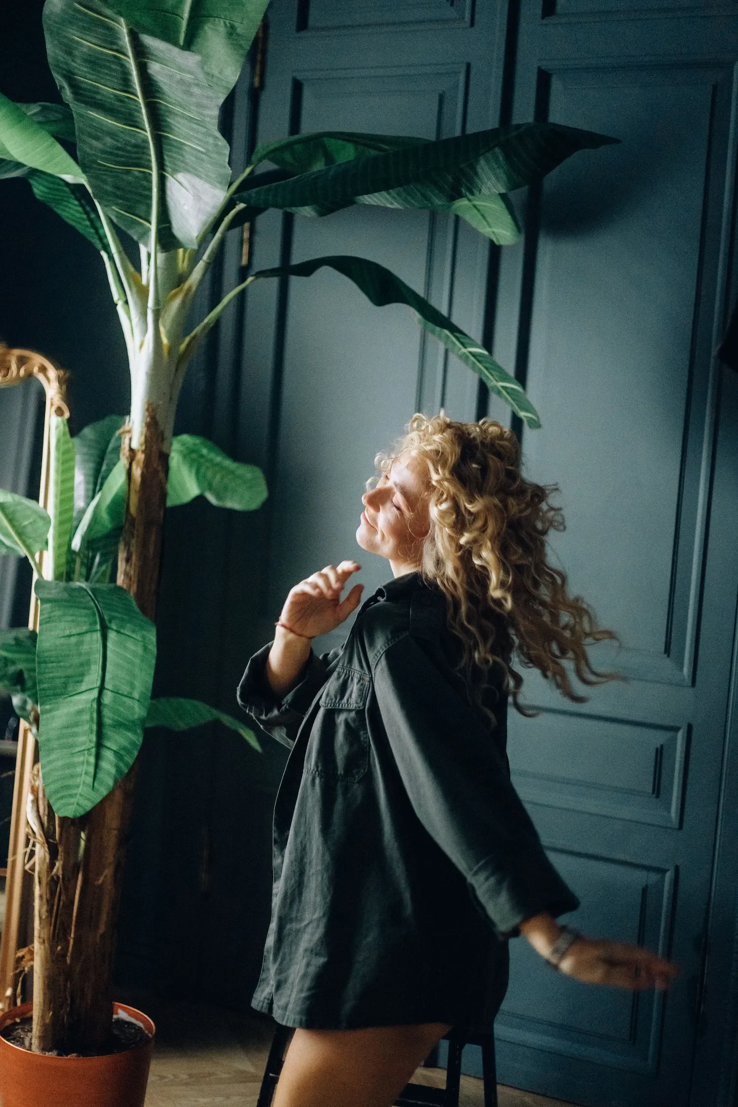 Une femme aux cheveux bouclés et roux se tient à côté d'une grande plante verte dans une pièce avec des murs bleus. Elle porte un manteau noir et une jupe courte, et semble se détendre ou apprécier un moment dans un intérieur sophistiqué.