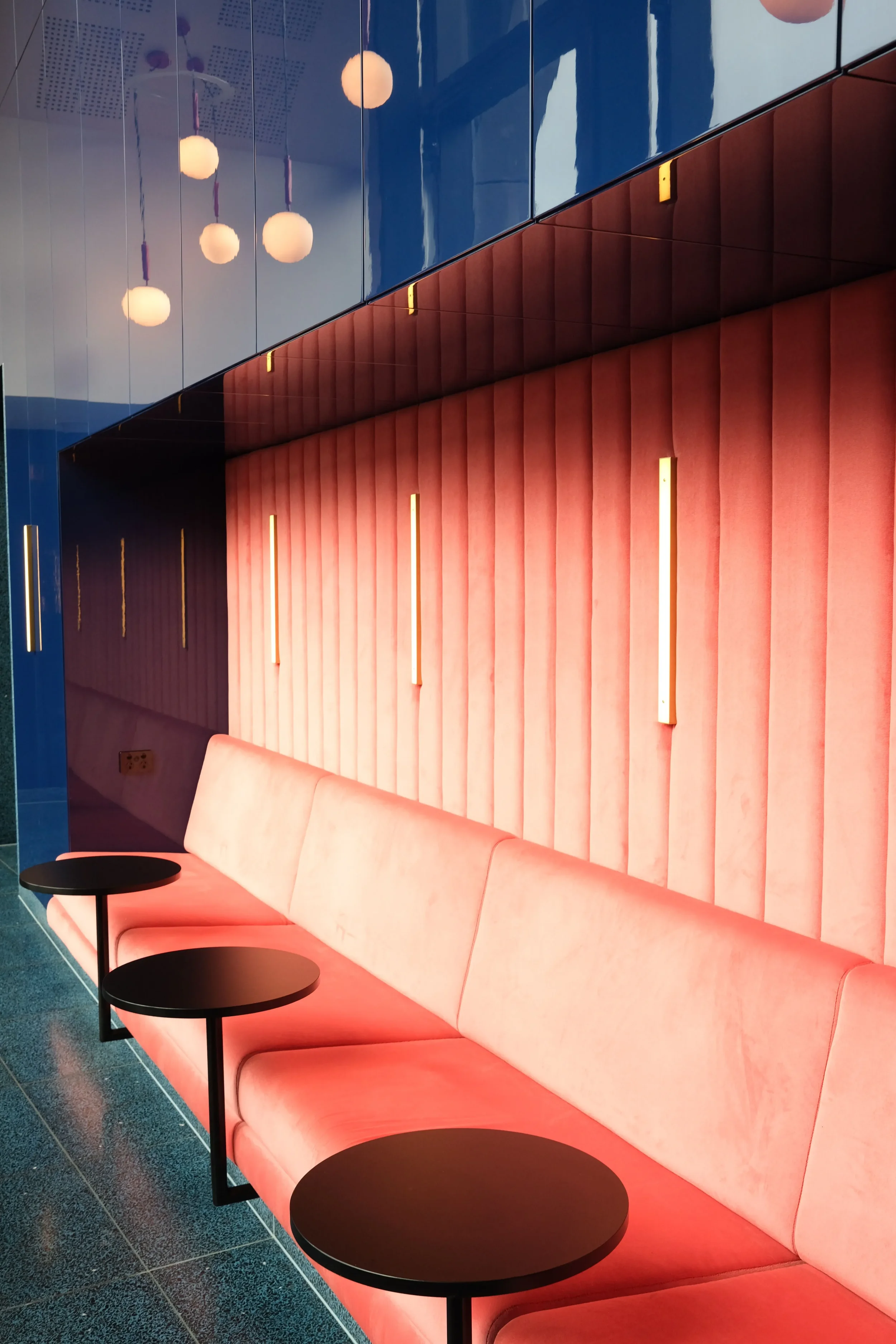 Une salle moderne avec un mur en velours rose avec des luminaires modernes, un canapé en velours rose, des tables rondes noires et un plafond avec des lampes sphériques