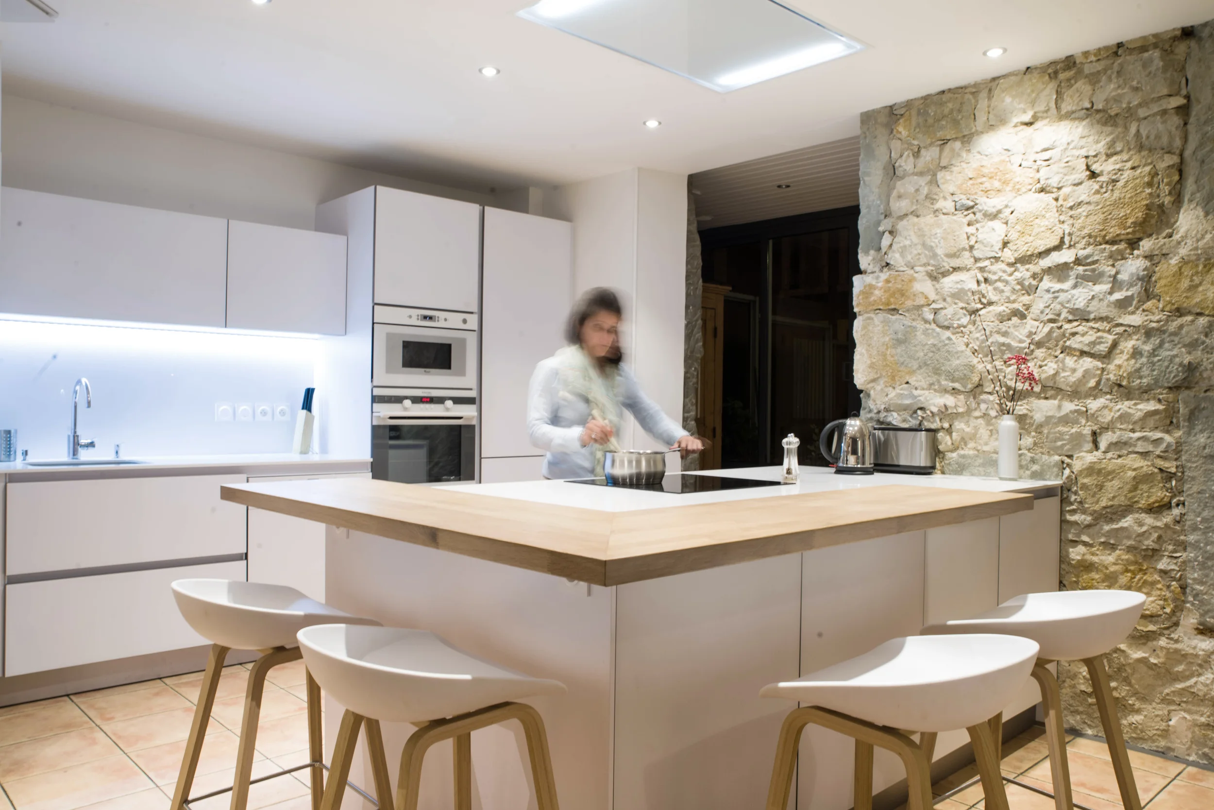 Une cuisine moderne lumineuse et chaleureuse avec un plan de travail en bois, des teintes blanches et un mur en pierre d'époque.