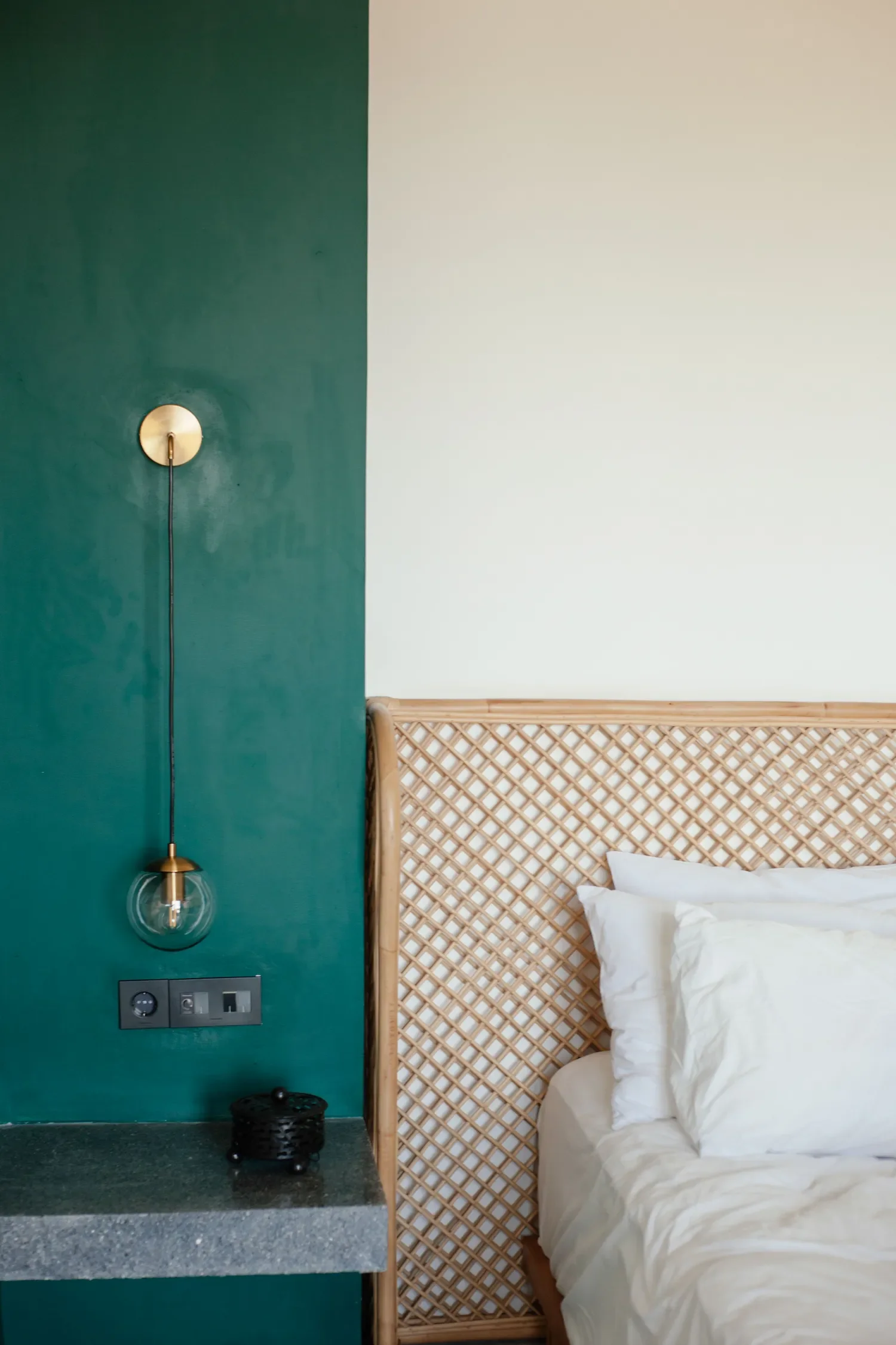 Chambre avec tête de lit en cannage, paroi verte avec lampe suspendue, et côté droit un lit avec draps blancs.