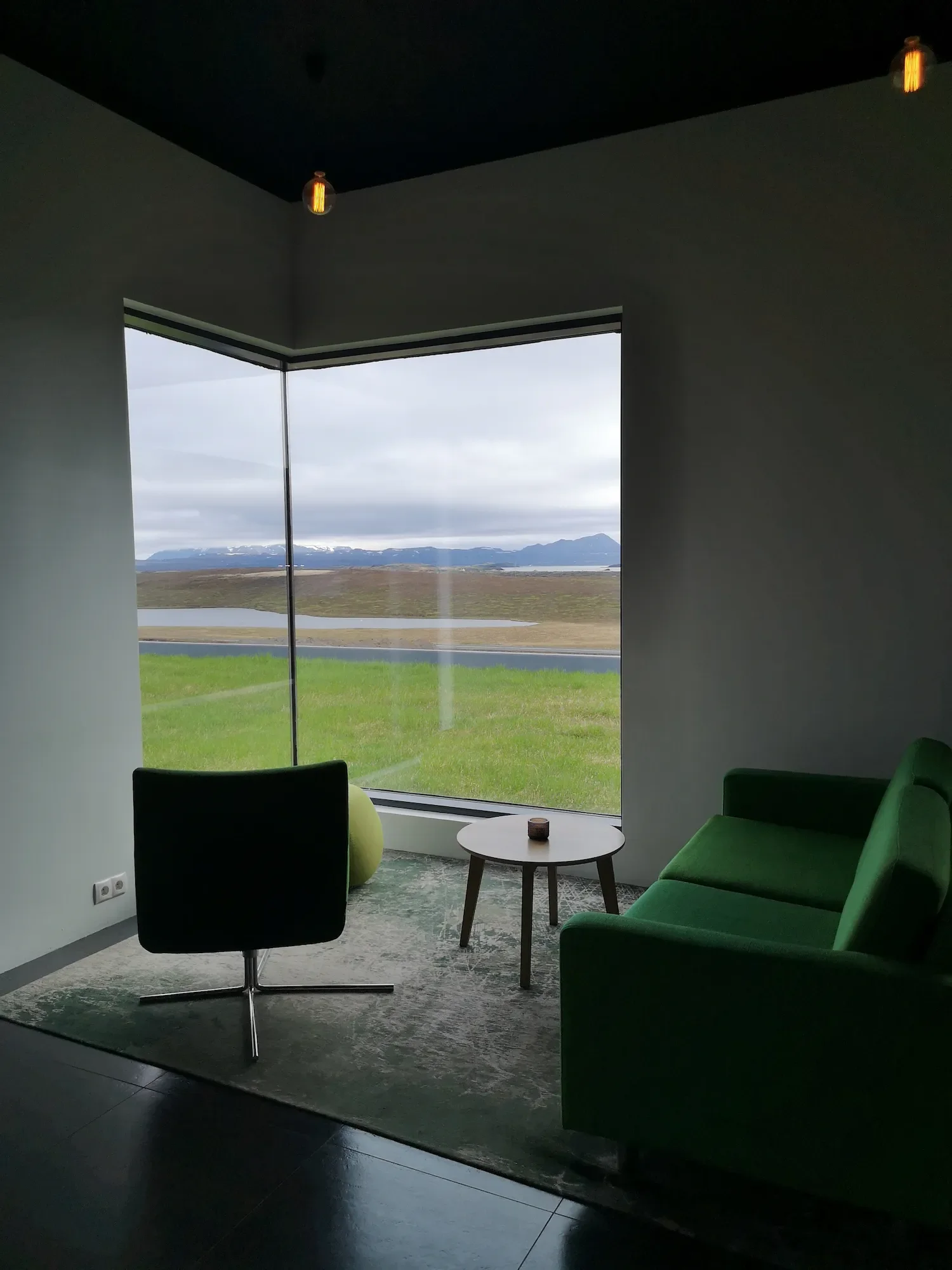 Intérieur d'une zone de repos dans le sein d'un hôtel islandais en pleine nature avec un canapé vert, une chaise noire, une petite table ronde, vue sur un paysage de montagnes et de plaines à travers une grande fenêtre. Quand la nature s'invite.