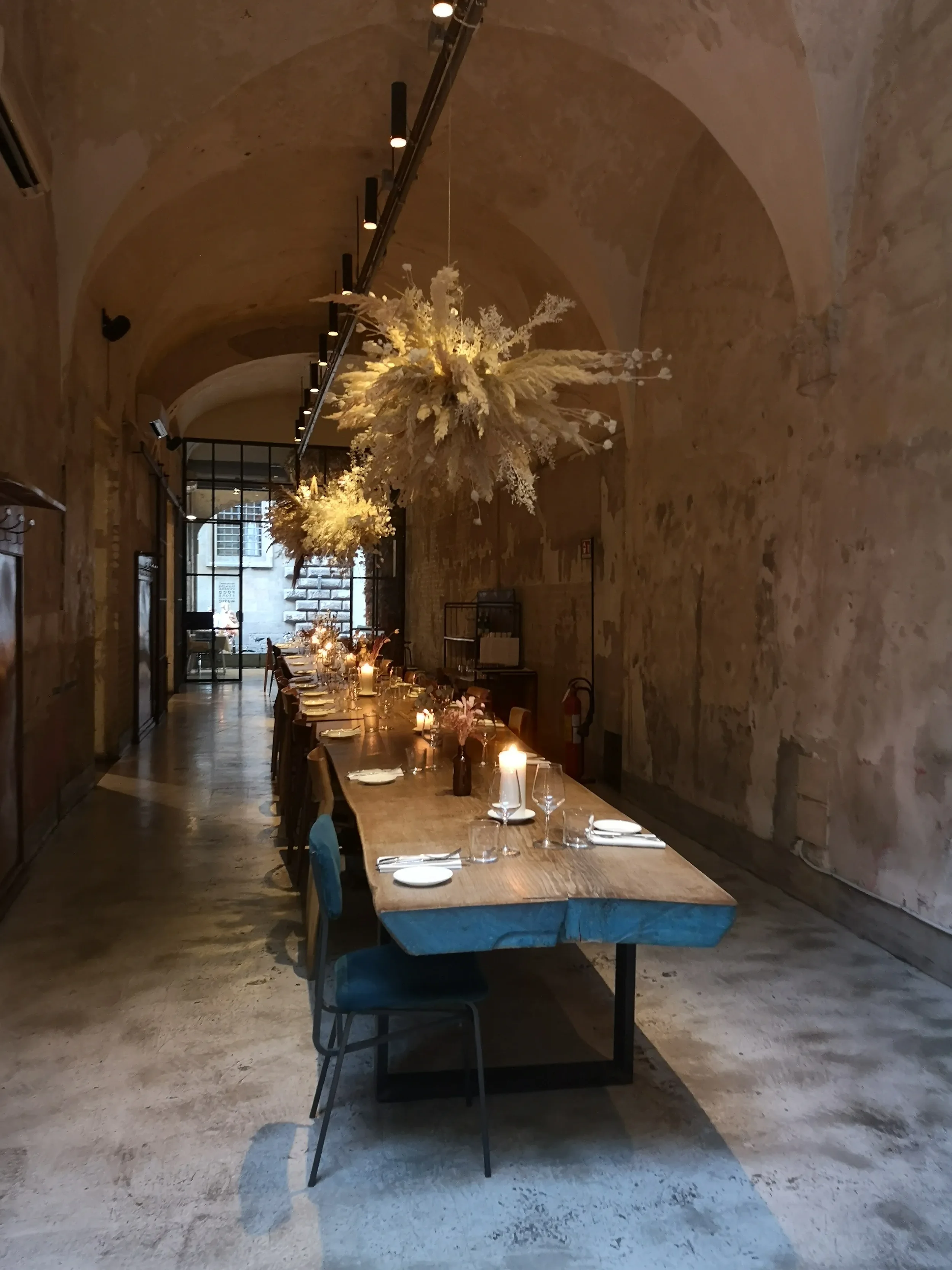 Une salle de restaurant constituée des matériaux bruts avec une longue table dressée pour un repas, décorée de bougies et de vases avec des fleurs séchées, sous un plafond voûté avec des suspensions florales.