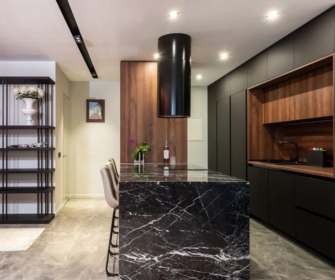 Cuisine moderne avec îlot en marbre noir, colonnes en bois, contraste entre un agencement sur mesure noir et bois, éclairages encastrés.