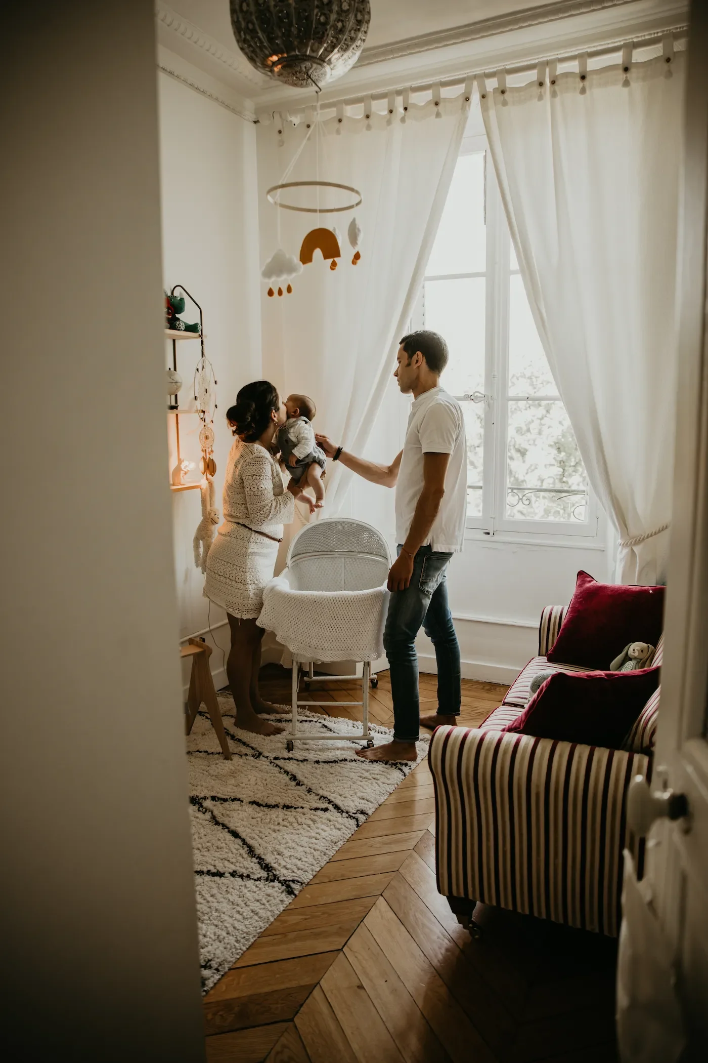 Chambre lumineuse et chaleureuse dans un appartement de style haussmannien. L'ambiance est intime, joyeuse et cosy avec des rideaux blancs, un tapis berbère, un berceau d'époque et un canapé à rayures rouges et blanches avec un coussin fuchsia.