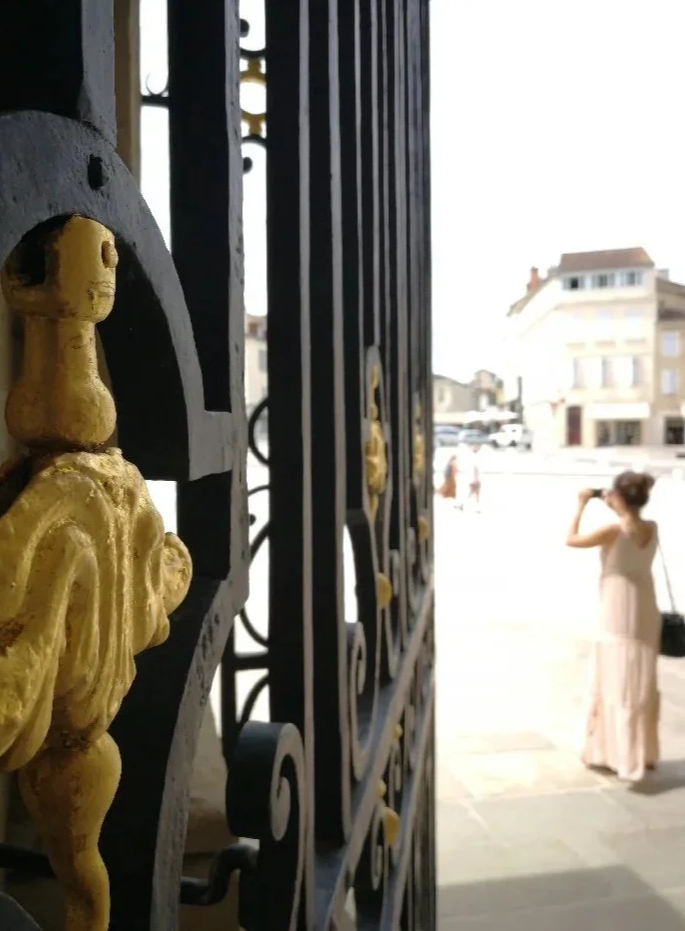 Une vue partielle d'une clôture noire avec des décorations dorées en forme de figures humaines, en premier plan. En arrière-plan, une femme portant une robe longue claire, tenant une caméra ou un téléphone, se tenant devant une architecture urbaine.