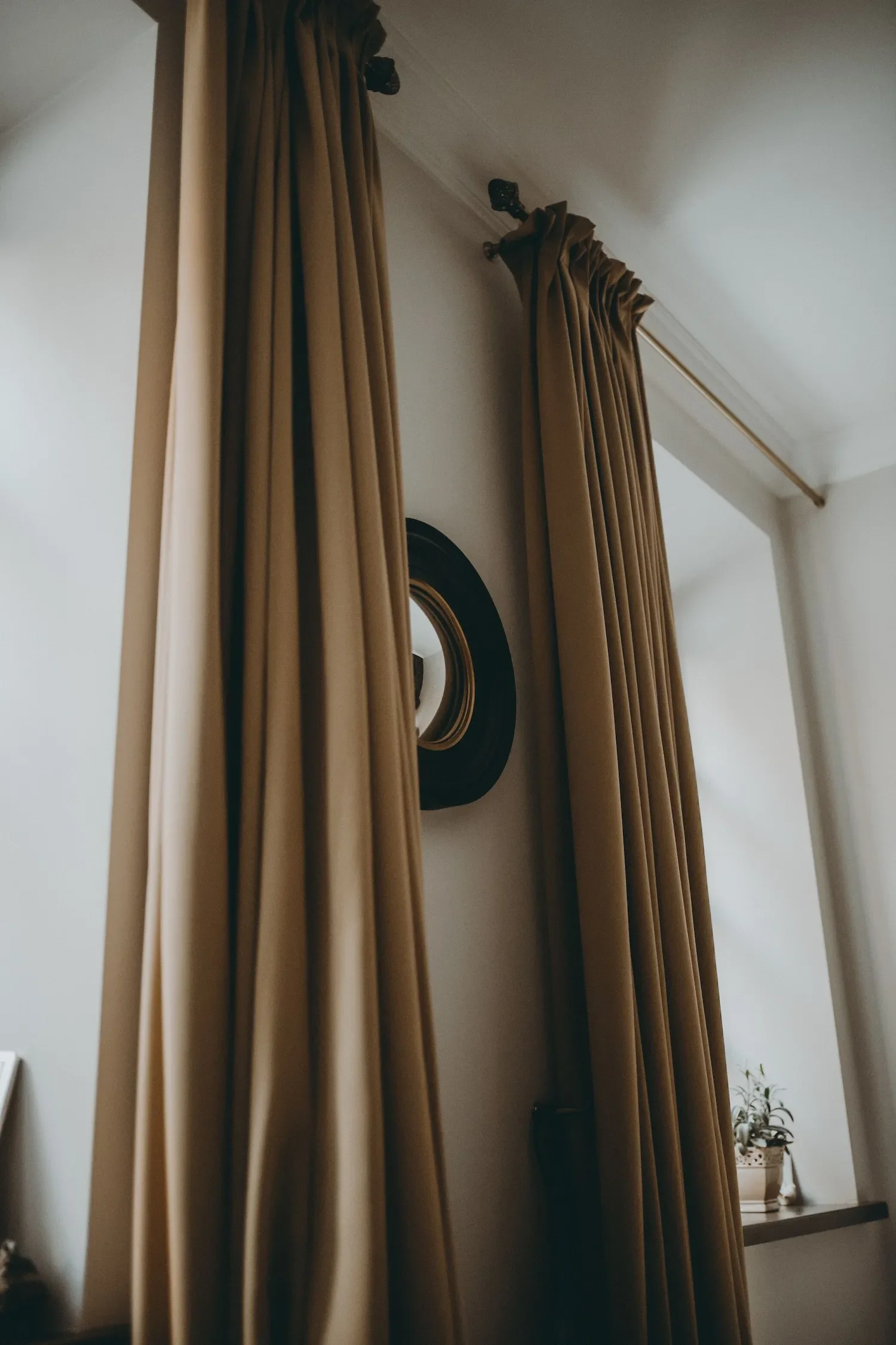 Une pièce intérieure lumineuse avec des rideaux marrons devant une fenêtre, un miroir convexe accroché au mur, pour sublimer la belle hauteur sous plafond.
