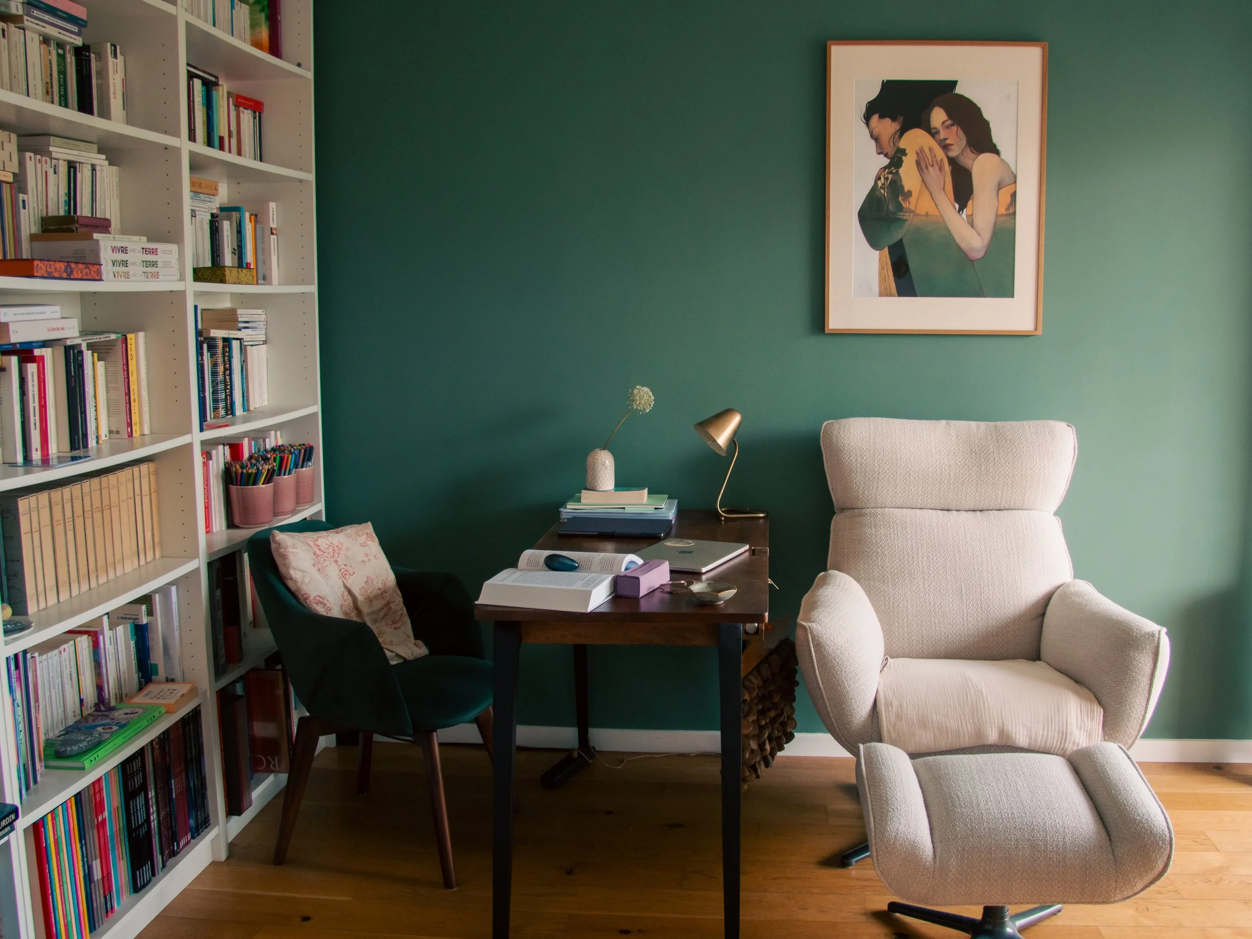 Un bureau avec une étagère remplie de livres, une table avec un ordinateur portable, une lampe, une plante, une chaise jaune et une grande peinture représentant deux femmes.