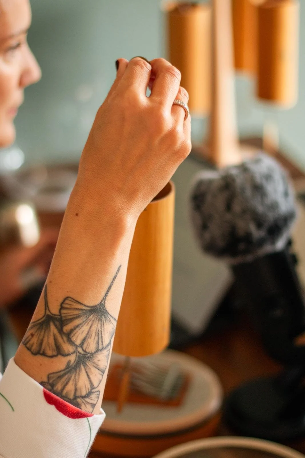 Première personne avec tatouage de branches de ginkgo sur le bras, tenant une petite chose entre le pouce et l'index, dans un intérieur avec lampes en bois et objet en pierre en arrière-plan.