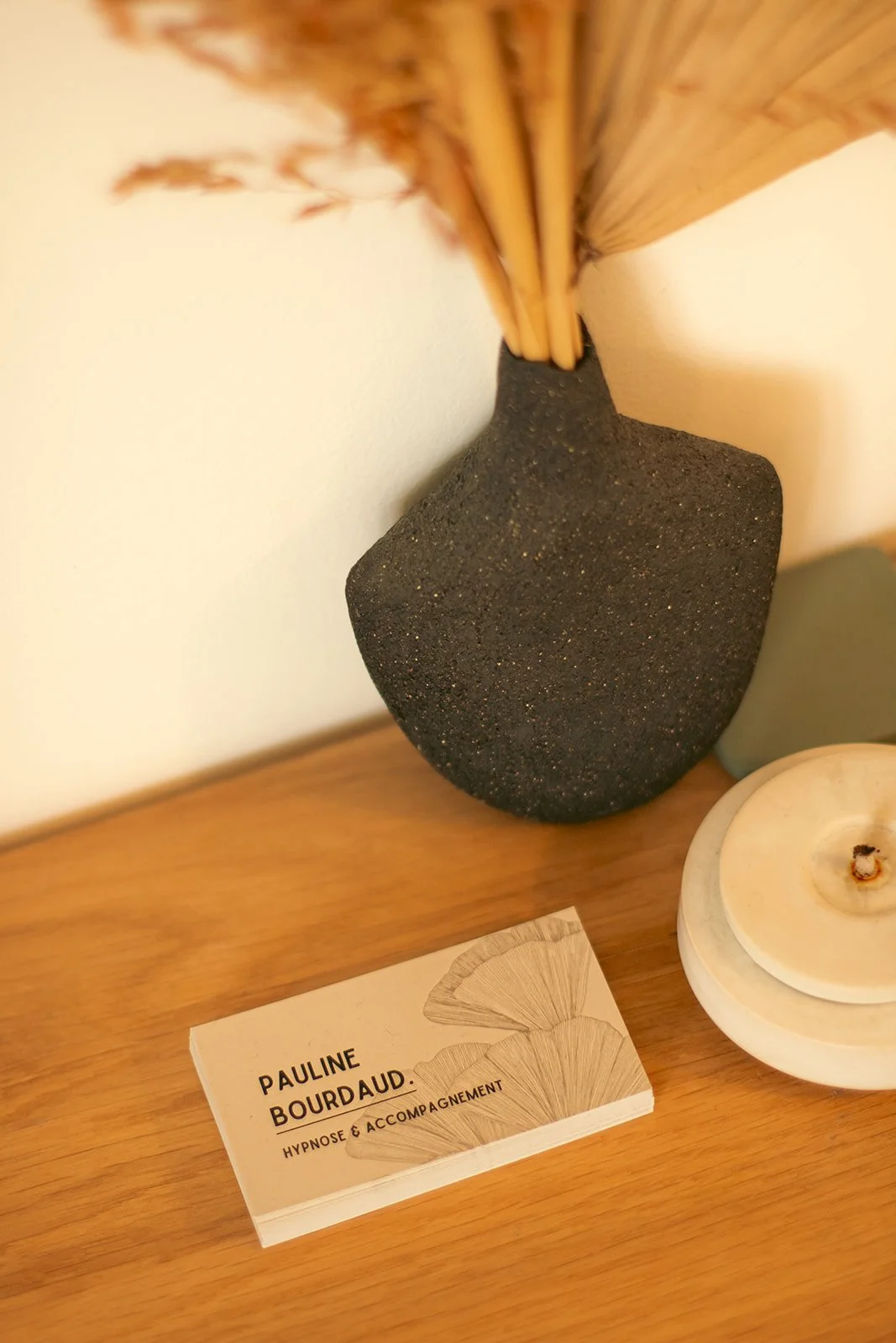 Une carte de visite beige avec le nom 'Pauline Bourdaud', mentionnant 'Hypnose & Accompagnement', posée sur une table en bois, avec un vase en pierre noire contenant des bâtons ou des branches en haut de l'image.