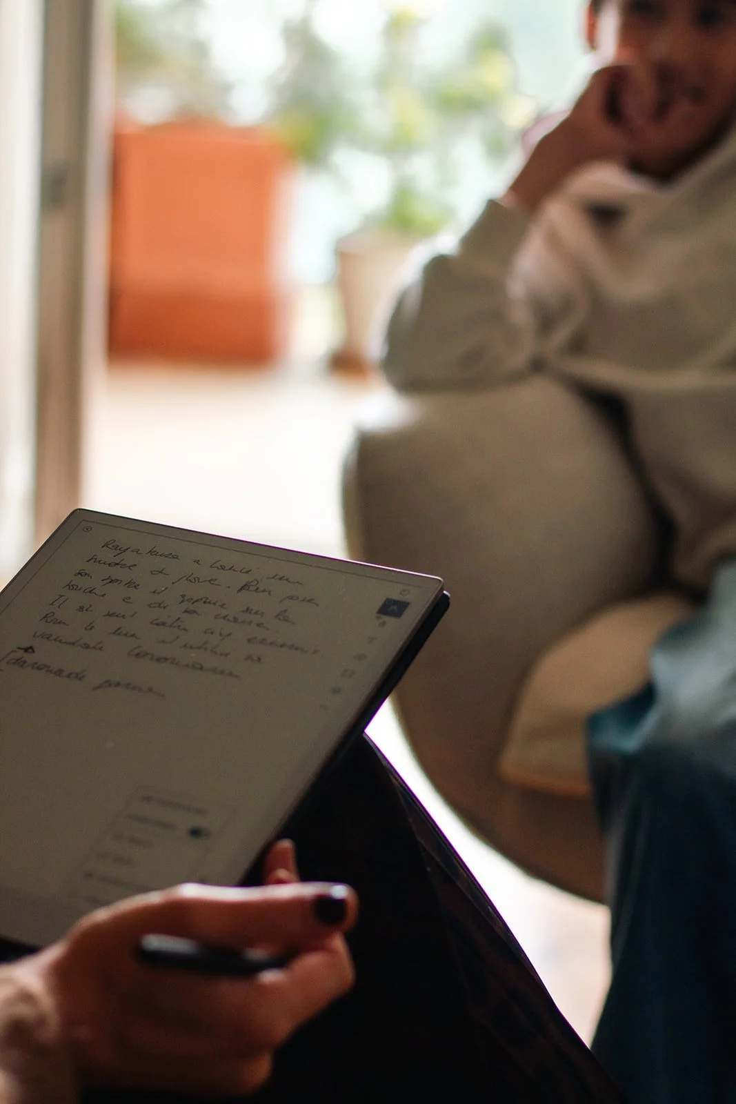 Une personne au premier plan tient un stylo et utilise une tablette pour prendre des notes manuscrites en français. Un autre personne, assise dans le fond, est visible partiellement et semble écouter ou discuter.