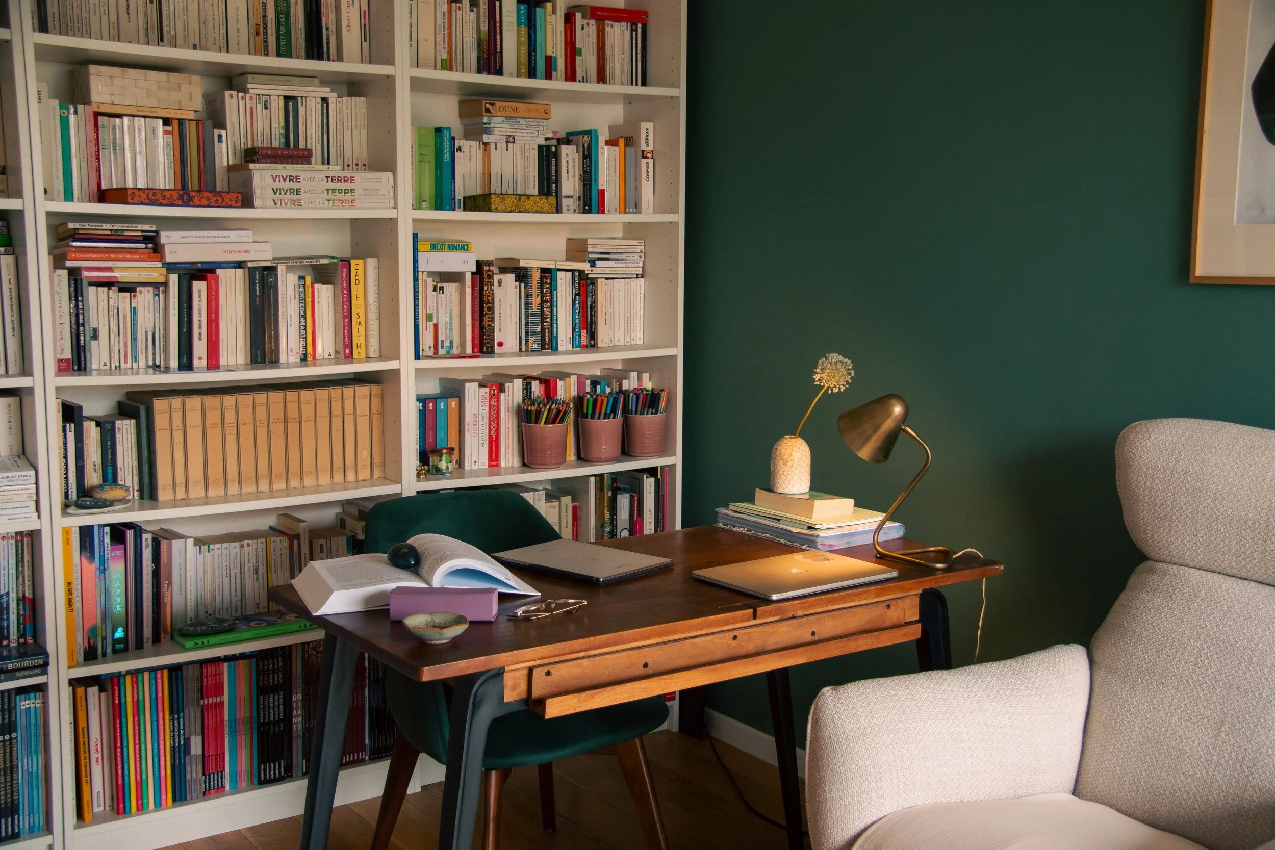 Une étagère blanche pleine de livres de différentes tailles et couleurs dans un salon avec un mur vert foncé. Sur la table en bois, il y a un ordinateur portable, un livre ouvert, un cahier, une lampe dorée avec un bras flexible et quelques petits objets. Il y a aussi une canette verte et une assiette à côté d'une chaise verte.