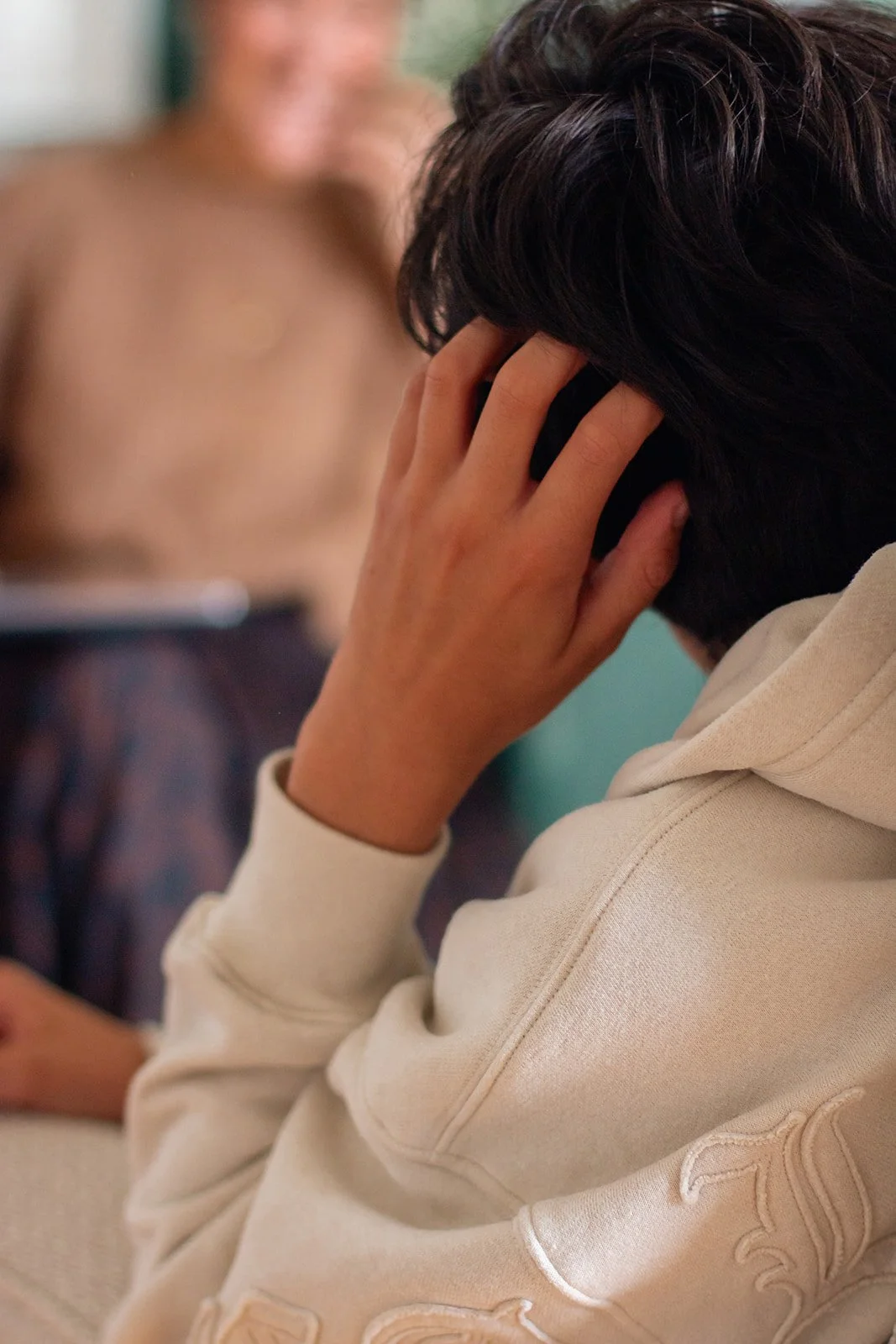 Une personne avec des cheveux foncés, portant un sweat beige, tient une oreillette ou un écouteur à l'oreille, avec une autre personne floue en arrière-plan.